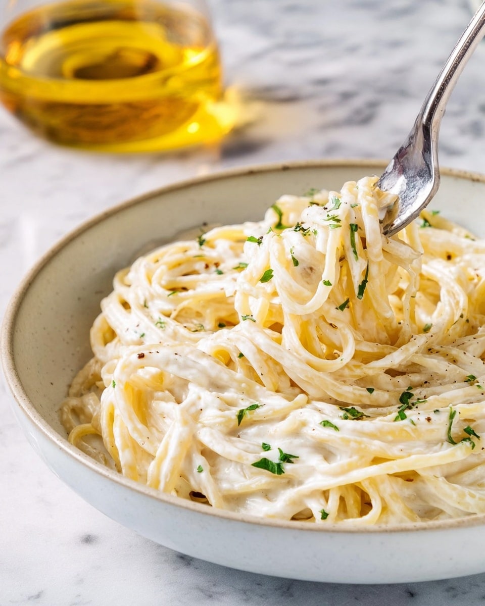 A bowl of creamy pasta with thick, smooth white sauce coating long, twisted spaghetti noodles. The pasta is lightly sprinkled with small pieces of fresh green herbs, adding a touch of color. A shiny silver fork is stuck into the pasta, lifting some strands in a soft spiral. The bowl is white and sits on a white marbled surface. In the background, a clear glass container with golden liquid is slightly blurred. photo taken with an iphone --ar 4:5 --v 7