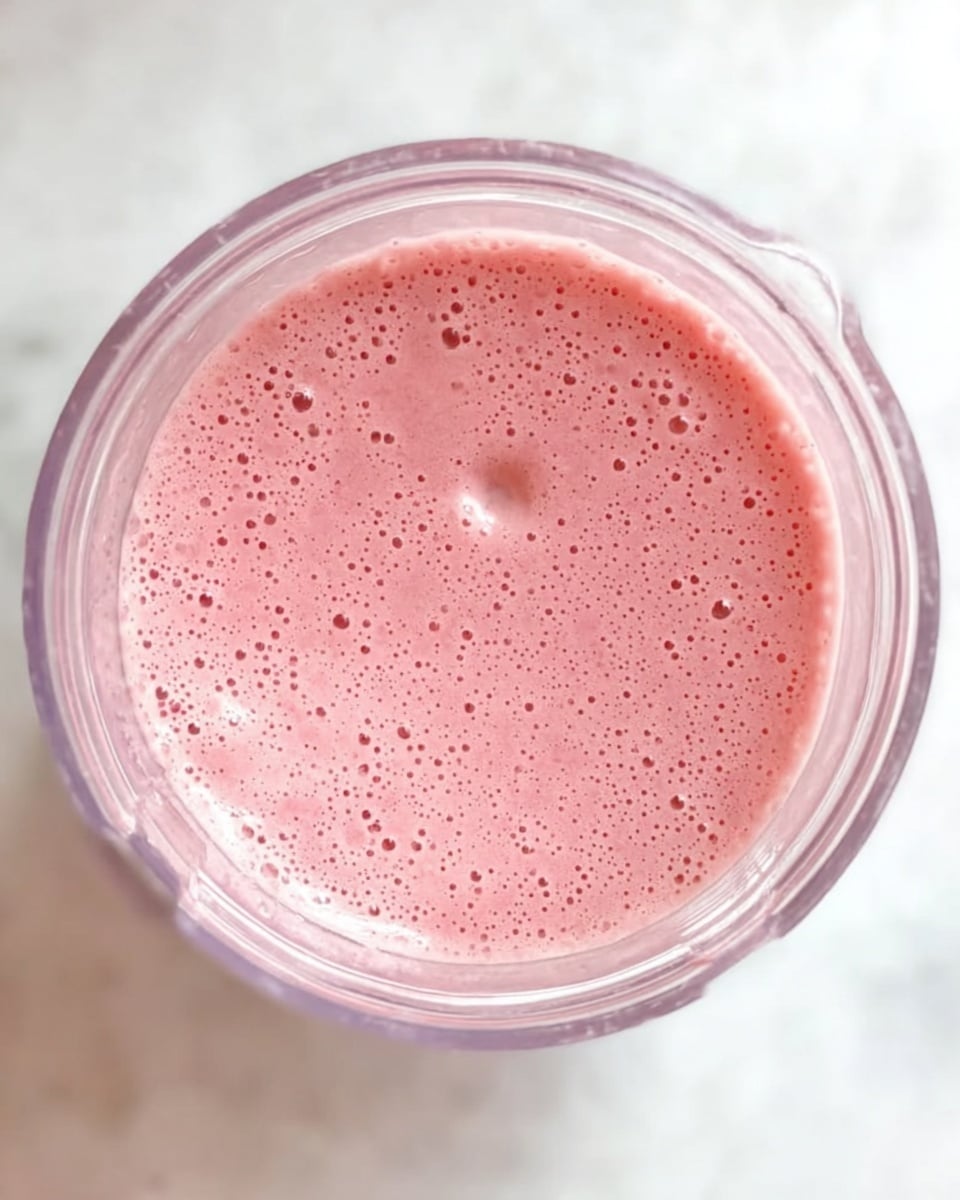 The image shows a clear blender jar filled with a single layer of pink smoothie that has many small bubbles and a few larger bubbles on its surface, giving it a light and airy texture. The blender sits on a white marbled surface, and the photo is taken from above showing the round shape of the jar’s top edge. photo taken with an iphone --ar 4:5 --v 7