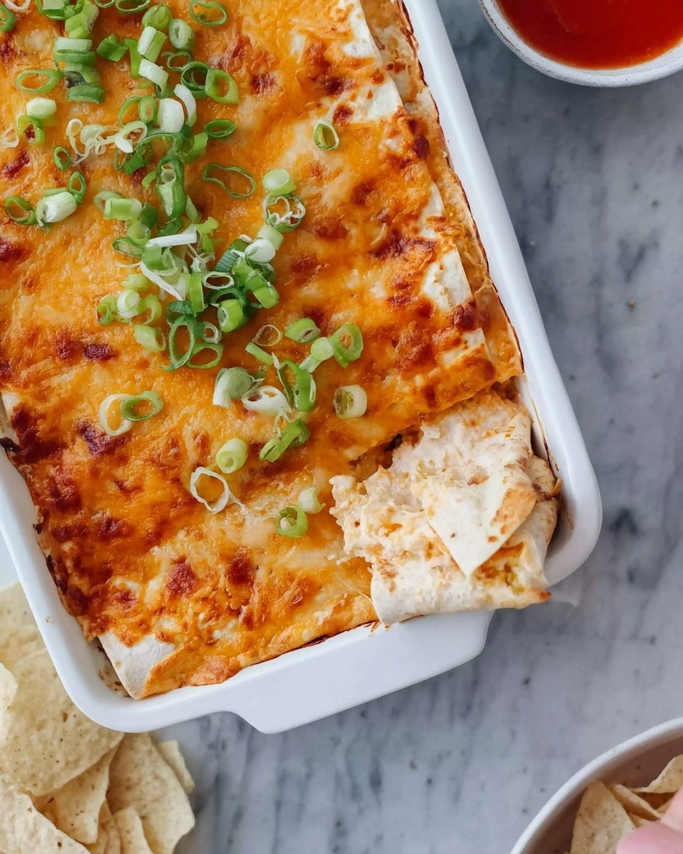 A close-up view of a white rectangular baking dish filled with layered enchiladas. The top layer is golden-brown melted cheese with some browned spots, decorated with thinly sliced green onions scattered over one corner. Below the cheese, soft pale tortillas wrapped around a filling that looks creamy and slightly orange. A woman's hand is lifting one rolled tortilla from the dish, showing the inside layers. Nearby, there are light beige tortilla chips on a white marbled surface, and part of a small white bowl with red sauce is visible. Photo taken with an iphone --ar 4:5 --v 7