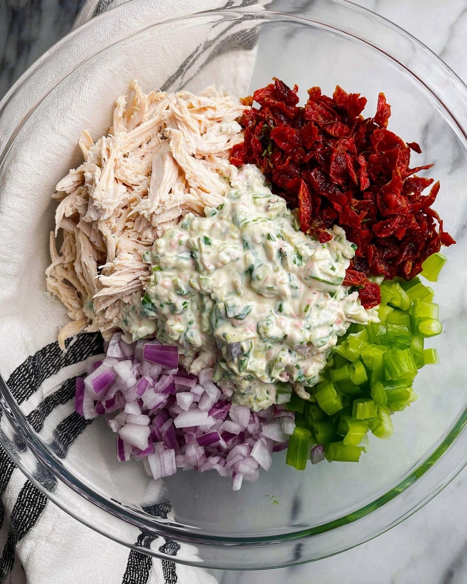 In a clear glass bowl, there are five visible layers arranged separately without mixing: top left pile of shredded light pink chicken, center a creamy white sauce with green herb bits, right a heap of finely chopped dark red sun-dried tomatoes, bottom left a pile of small purple onion pieces, and bottom right a pile of small green celery pieces. The bowl sits on a white marbled surface with part of a white and black cloth visible under the bowl's edge. Photo taken with an iphone --ar 4:5 --v 7