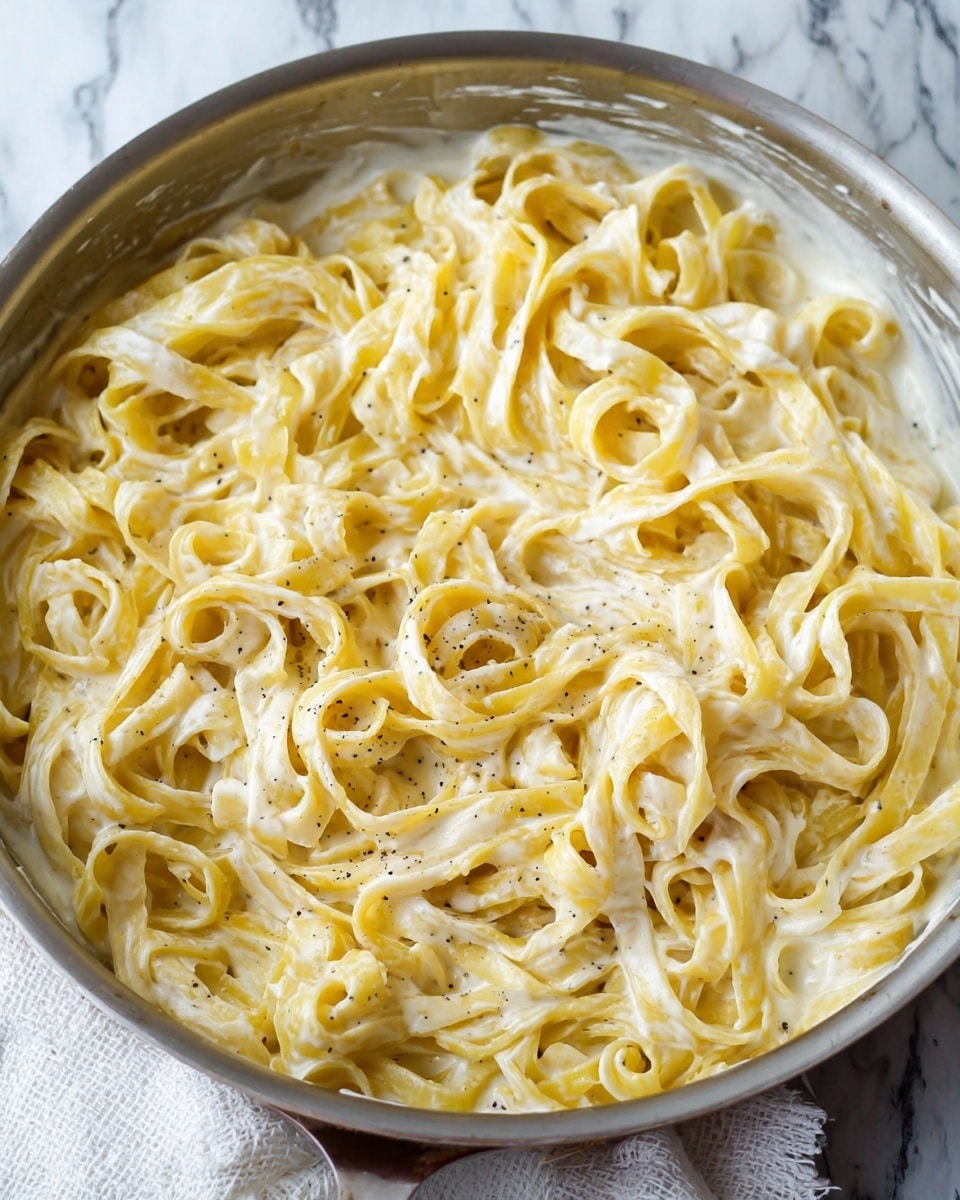 A close-up view of a large metal pan filled with creamy fettuccine pasta, showing one thick layer of wide, flat yellow noodles covered in a smooth, white creamy sauce with small black pepper specks evenly spread. The pasta strands are somewhat twisted and shiny, giving a rich, soft texture look. The pan sits on a white marbled surface with a glimpse of a white cloth underneath. Photo taken with an iphone --ar 4:5 --v 7