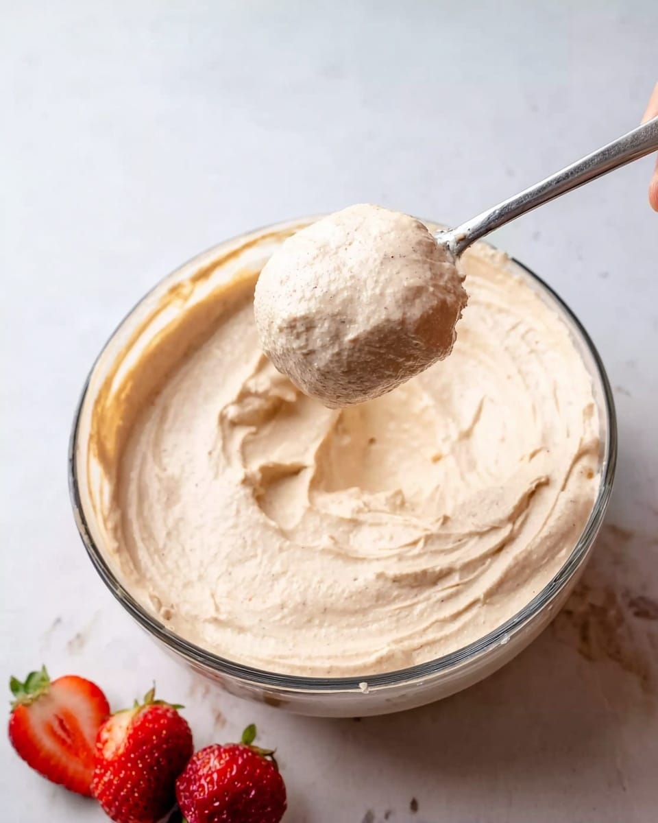 The image shows a close-up of a large clear glass bowl filled with creamy, light beige dip or spread. A silver spoon is held above the bowl by a woman's hand, scooping a generous amount of the smooth, thick dip. Around the bowl, there are a few pieces of red strawberries resting on the white marbled surface, adding a pop of color. The background is simple and bright, highlighting the dip's texture and the fresh fruit. Photo taken with an iphone --ar 4:5 --v 7
