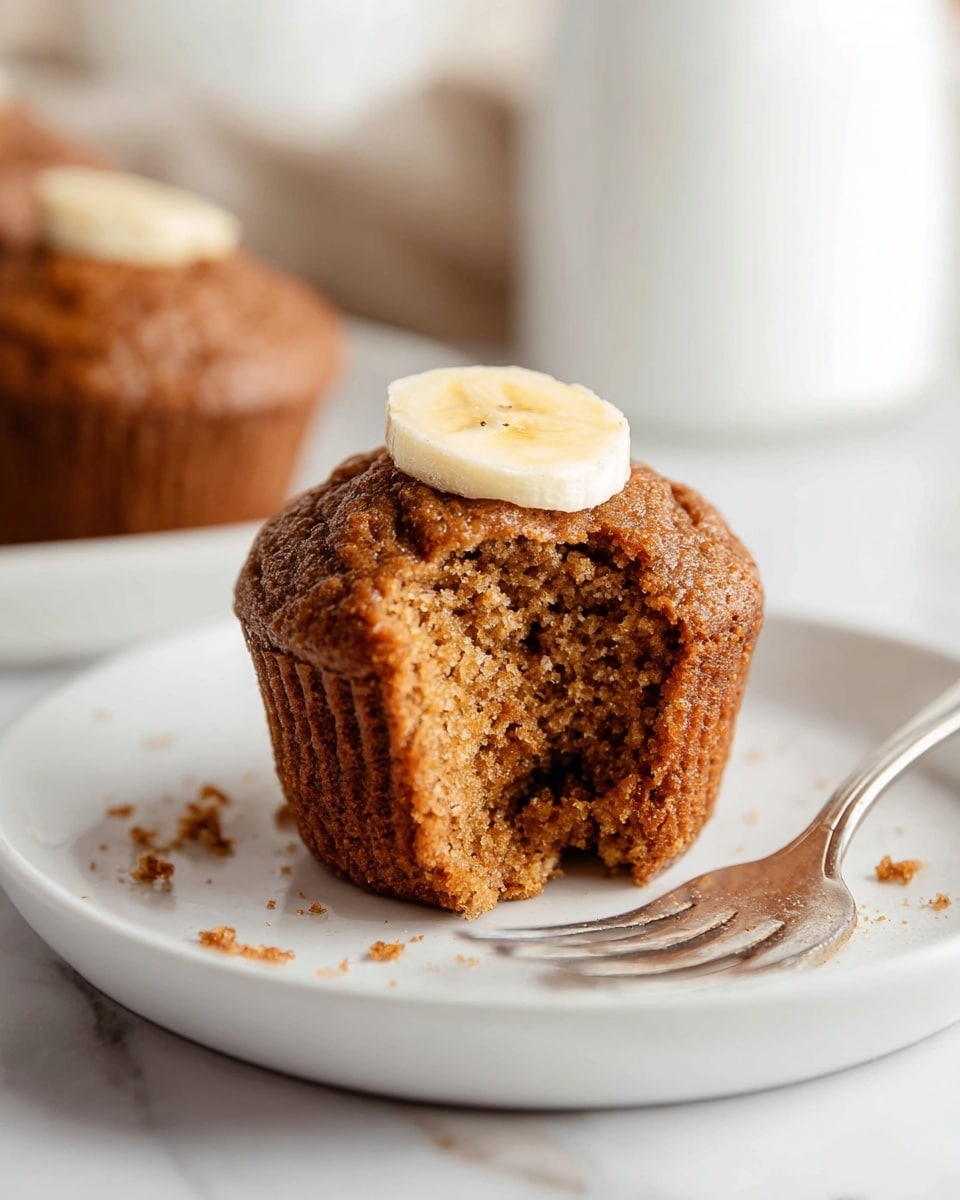 A single brown muffin with a soft, crumbly texture sits on a white plate, showing a large bite taken from its front side revealing its moist interior. It has a small round slice of banana placed flat on top as a simple decoration. Behind the muffin, a silver fork lies on the plate with some crumbs on it. The surface beneath is white marble, and the background is softly blurred with another muffin partly visible. The light is natural and bright, highlighting the muffin’s warm tones. Photo taken with an iphone --ar 4:5 --v 7