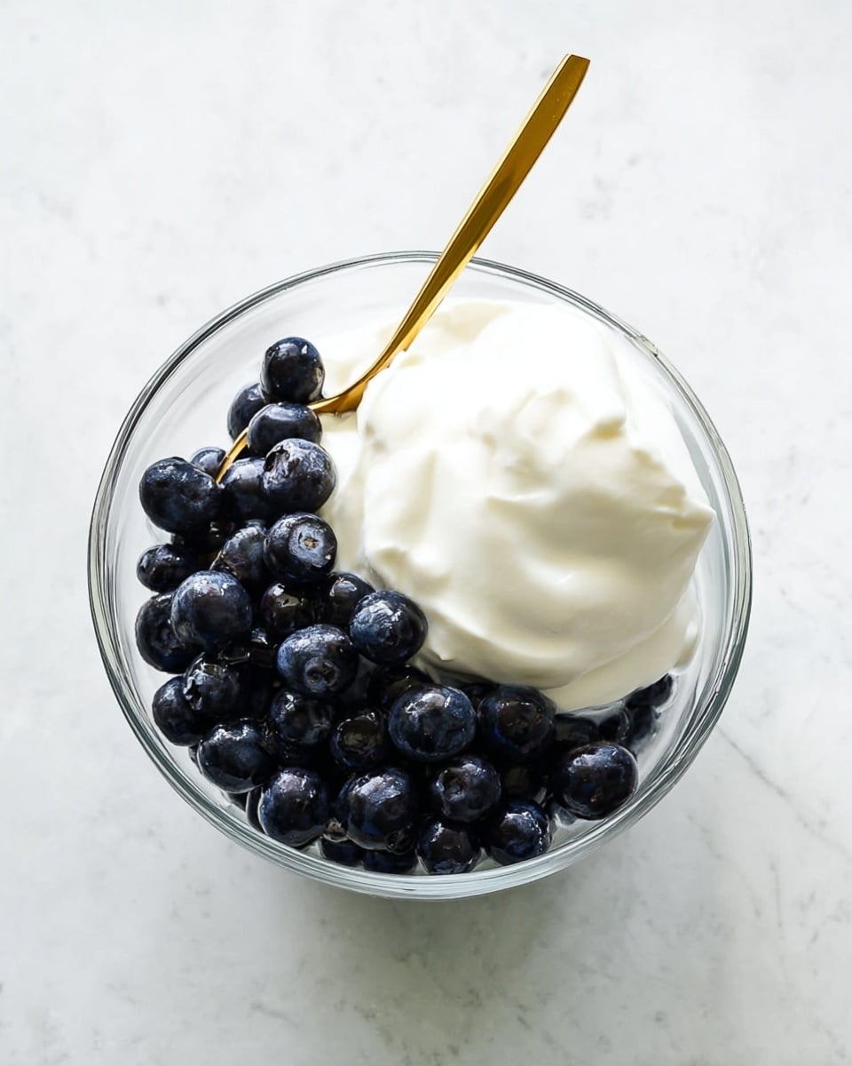 A clear glass bowl holds two main layers: a bottom layer of dark blue, round blueberries filling about two-thirds of the bowl, topped with a dollop of thick, smooth white yogurt sitting mostly on one side. A gold spoon with a slim handle is placed in the bowl, leaning on the yogurt and resting slightly on the blueberries underneath. The bowl sits on a white marbled surface. photo taken with an iphone --ar 4:5 --v 7
