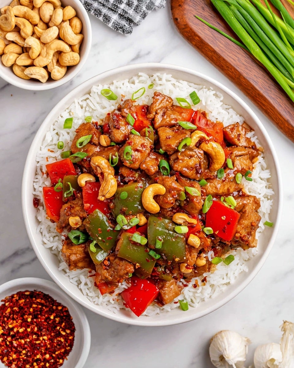 A white bowl is filled with a base layer of white rice with separate grains. On top, there is a thick layer of cooked meat pieces in a glossy brown sauce, mixed with large, bright red and green bell pepper chunks and whole golden cashews. The dish is sprinkled with chopped green onions and tiny red chili flakes, adding color contrast. Around the bowl, there is a small white bowl of whole cashews, a wooden cutting board with green onions and a garlic bulb, and a small white bowl filled with red chili flakes, all placed on a white marbled surface. photo taken with an iphone --ar 4:5 --v 7