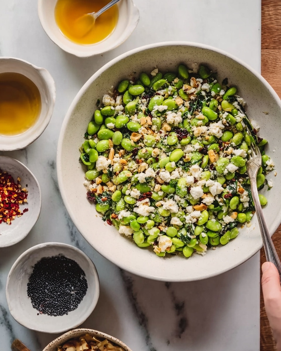 The image shows a large white bowl filled with a fresh salad made of bright green edamame beans, crumbled white cheese, and small pieces of light brown nuts. The salad is mixed with tiny black seeds and some dark green leafy bits, all spread evenly across the bowl. The bowl sits on a white marbled surface, next to small white bowls containing black seeds, red chili flakes, and a golden yellow liquid, with a spoon placed inside one bowl. A woman's hand is holding a fork over the edge of the salad bowl. The photo taken with an iphone --ar 4:5 --v 7