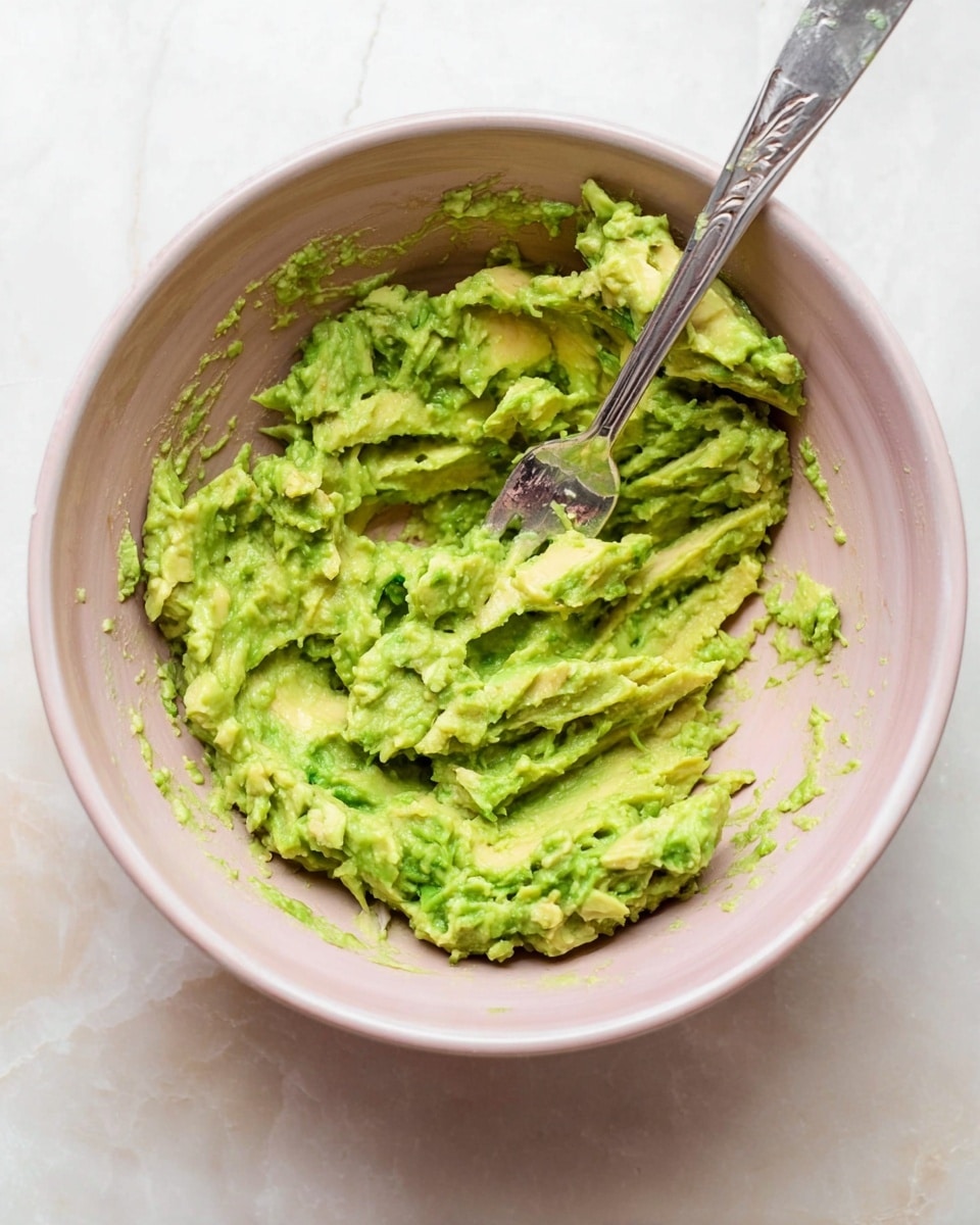 A close-up photo of pale pink bowl filled with mashed avocado that shows a rough texture with some chunks and wide fork marks pressing the avocado against the bowl. The mashed avocado is bright green with some lighter green and yellow-green areas. The silver fork is resting inside the bowl, with its handle leaning on the bowl's edge, and the photo is set against a white marbled surface. photo taken with an iphone --ar 4:5 --v 7