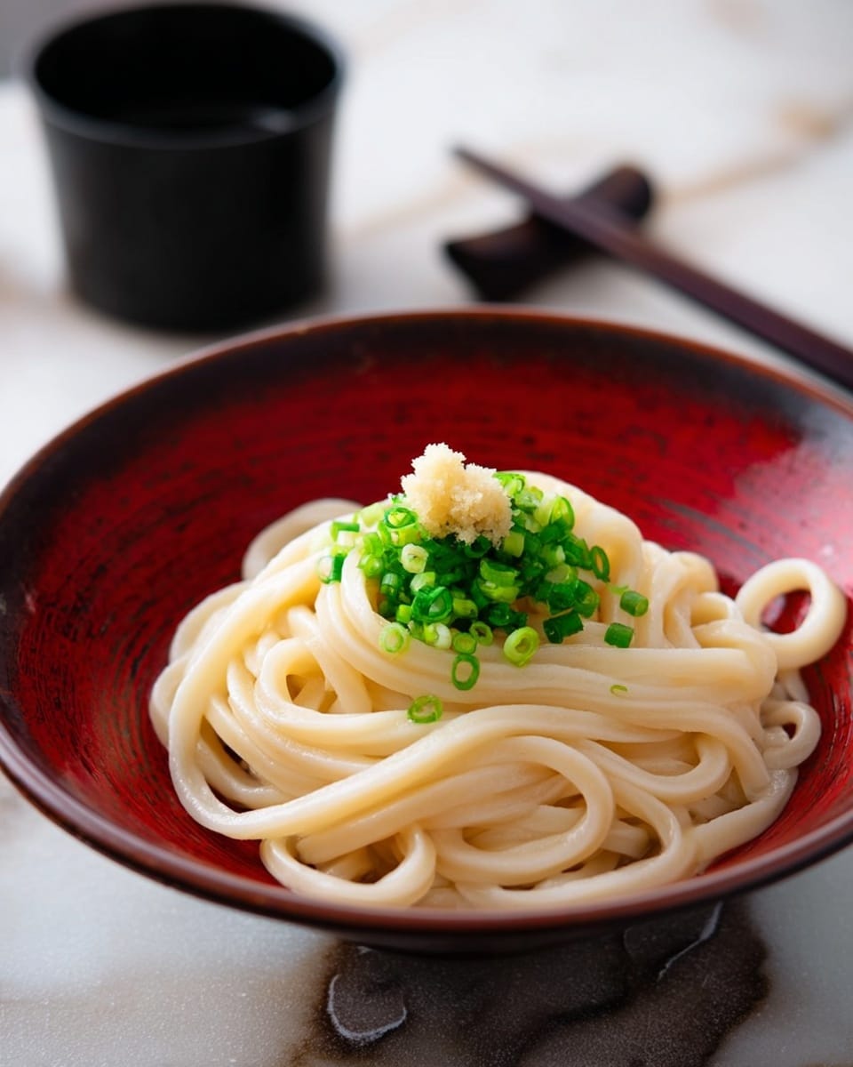 A white bowl holds a neat pile of thick, pale yellow udon noodles arranged in a loose mound. On top, there is a small heap of finely chopped bright green scallions, and resting above the scallions is a small dollop of light beige grated ginger. The bowl has a deep red interior with a slightly rough texture, contrasting with the smooth noodles. The scene is set on a white marbled surface with chopsticks and a black soy sauce container blurred in the background. Photo taken with an iphone --ar 4:5 --v 7
