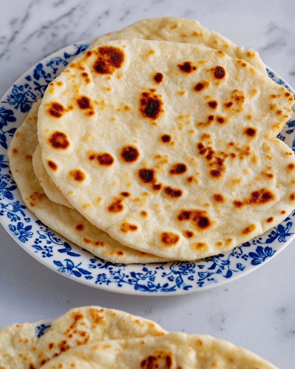 A single flat round bread with a light beige color is shown, covered with many small dark brown toasted spots all over its surface, indicating it is cooked. The bread has a soft texture with slight puffiness and some uneven edges. Part of a white plate with blue floral patterns is visible at the bottom, holding more of the same bread. The background is a white marbled surface. Photo taken with an iphone --ar 4:5 --v 7