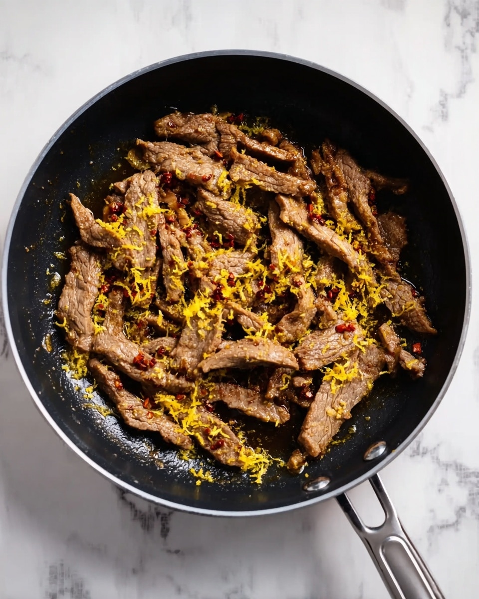 A black frying pan sits on a white marbled surface, filled with about one layer of cooked meat strips that have a brown, slightly crispy texture. The meat is sprinkled with small bits of red chili flakes and bright yellow lemon zest spread evenly on top, adding a vibrant contrast to the darker meat. The pan handle extends to the right side of the image. Photo taken with an iphone --ar 4:5 --v 7