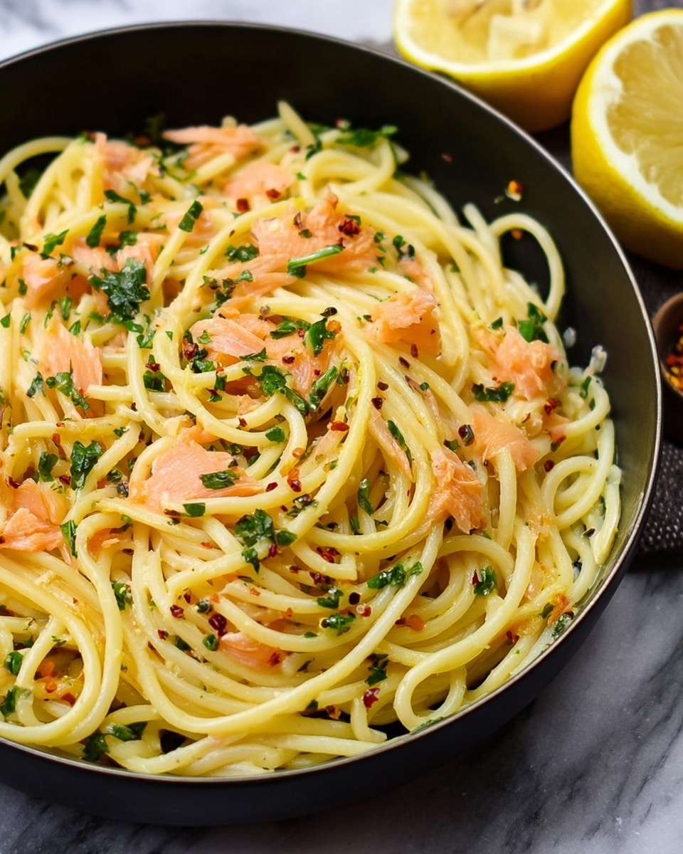 A close-up of a black bowl filled with light yellow spaghetti noodles that are loosely twirled and mixed with small pieces of pale orange smoked salmon. Green herbs and small red chili flakes are sprinkled evenly throughout the dish. The bowl sits on a surface with a white marbled texture, and behind the bowl are two lemon halves showing the bright yellow inside. The scene has a simple and fresh look with natural lighting. photo taken with an iphone --ar 4:5 --v 7