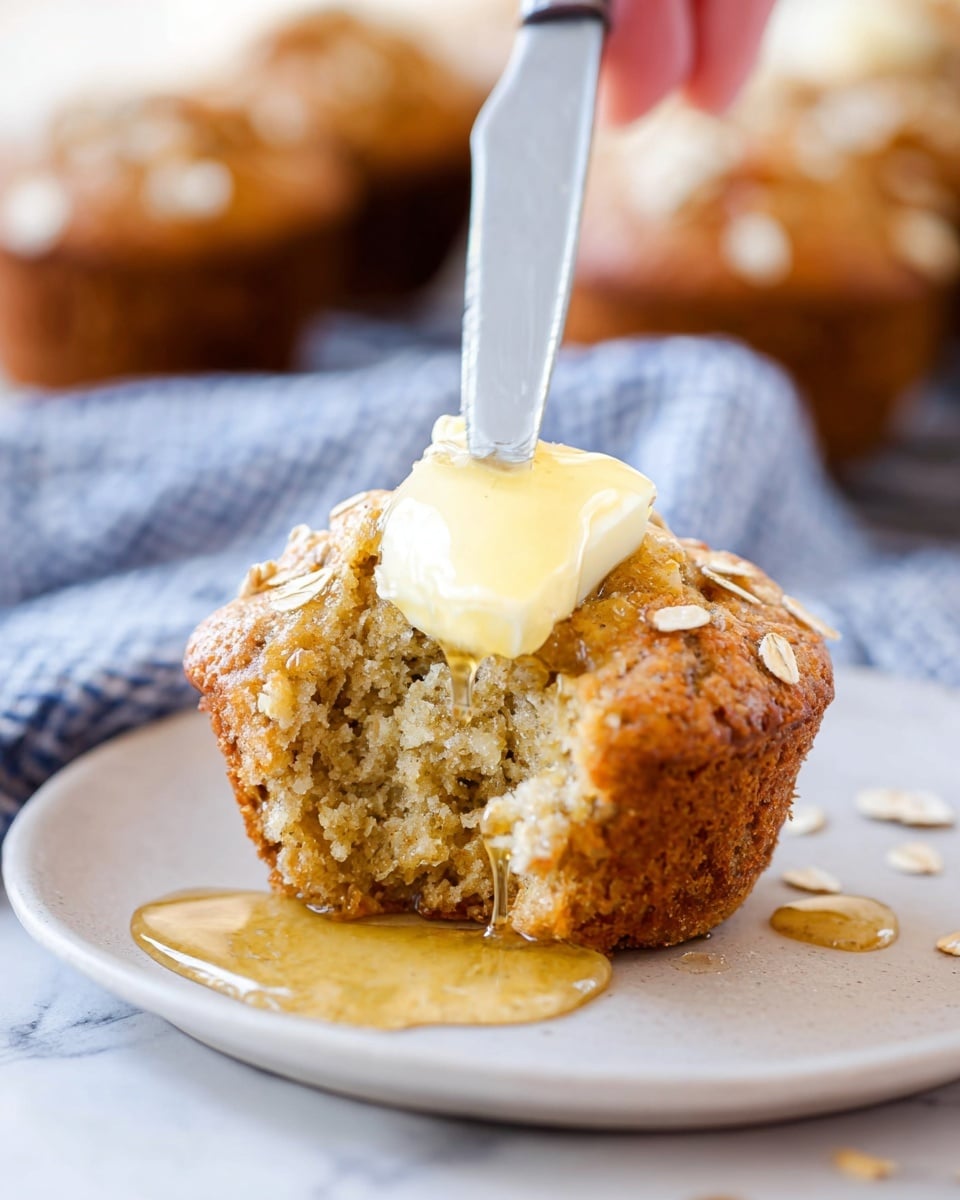 A close-up photo of a golden brown muffin with visible oats on top, placed on a white plate over a white marbled surface. The muffin has a bite taken out showing its soft, crumbly inside. A woman's hand holds a silver knife, spreading a small block of light yellow butter on top of the muffin. Honey is drizzled over the muffin and plate, creating shiny, sticky pools around the muffin base. In the background, there are blurred muffins on a blue and white checkered cloth. Photo taken with an iphone --ar 4:5 --v 7