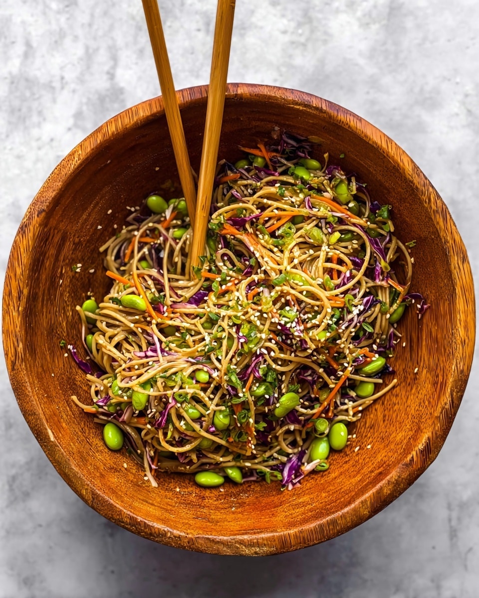 A large wooden bowl holds a colorful noodle salad with thin, light brown noodles swirling in the center, mixed with bright green edamame beans and thin strips of white and purple cabbage scattered throughout. Thin orange carrot strips and small green herbs are spread evenly across the dish, topped with a sprinkle of light beige sesame seeds. Two chopsticks are placed inside the bowl, one resting on the edge and the other standing upright amid the noodles. The bowl sits on a white marbled surface. Photo taken with an iphone --ar 4:5 --v 7