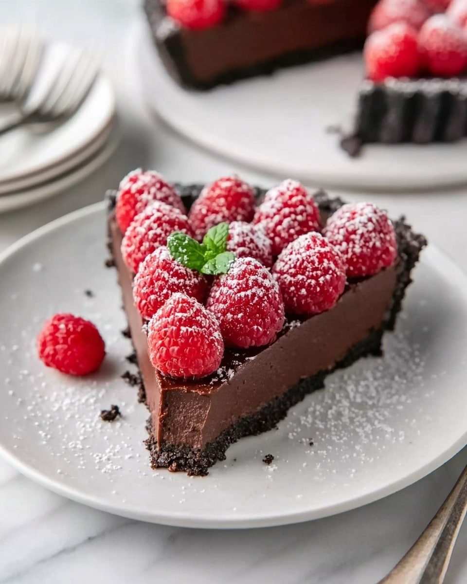 A slice of rich, dark chocolate tart is placed on a white plate, with a crumbly dark chocolate crust forming the bottom layer. On top of the crust is a thick, smooth layer of dark chocolate filling. The tart is topped with a neat row of bright red raspberries, dusted lightly with white powdered sugar, and garnished with a small green mint leaf in the center. The plate is resting on a white marbled surface, with another empty white plate and fork blurred in the background. A woman's fingers are gently touching the edge of the tart slice. Photo taken with an iphone --ar 4:5 --v 7