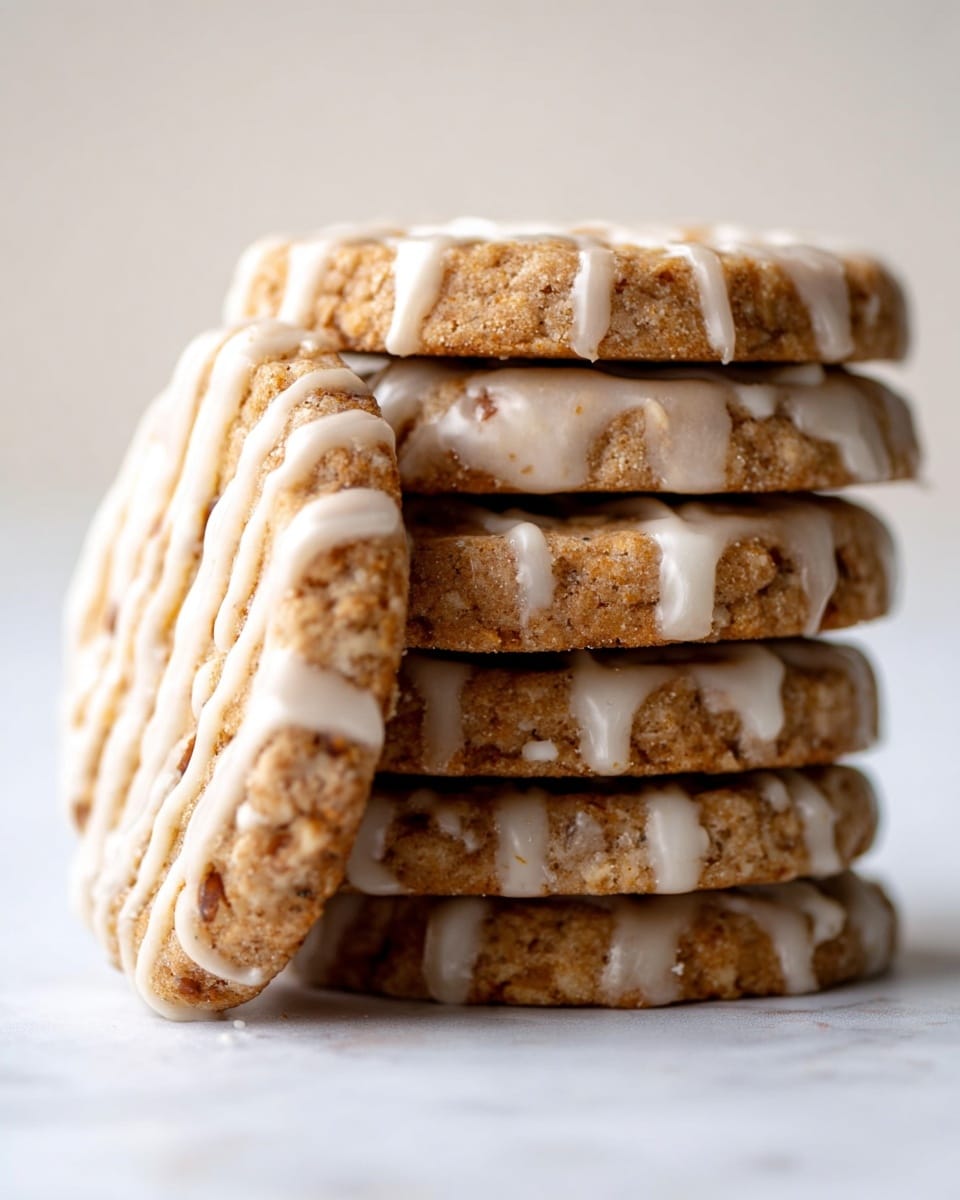 A close-up of a stack of four round cookies with a rough, nutty texture and light brown color. Each cookie has a scalloped edge and is drizzled with thin, white icing, some dripping slightly down the sides. The cookies stand vertically on a white marbled surface, with one cookie leaning against the stack on the left side. The background is softly blurred in light neutral tones. photo taken with an iphone --ar 4:5 --v 7