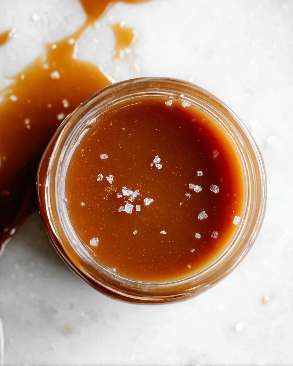A close-up top view of a small glass jar filled with smooth caramel sauce, with a shiny light brown surface. Tiny bits of white salt crystals are scattered on top of the sauce, adding texture. The jar is placed on a white marbled surface, with a bit of caramel spilled near the jar's edge. The caramel looks thick and glossy without any layers visible inside photo taken with an iphone --ar 4:5 --v 7