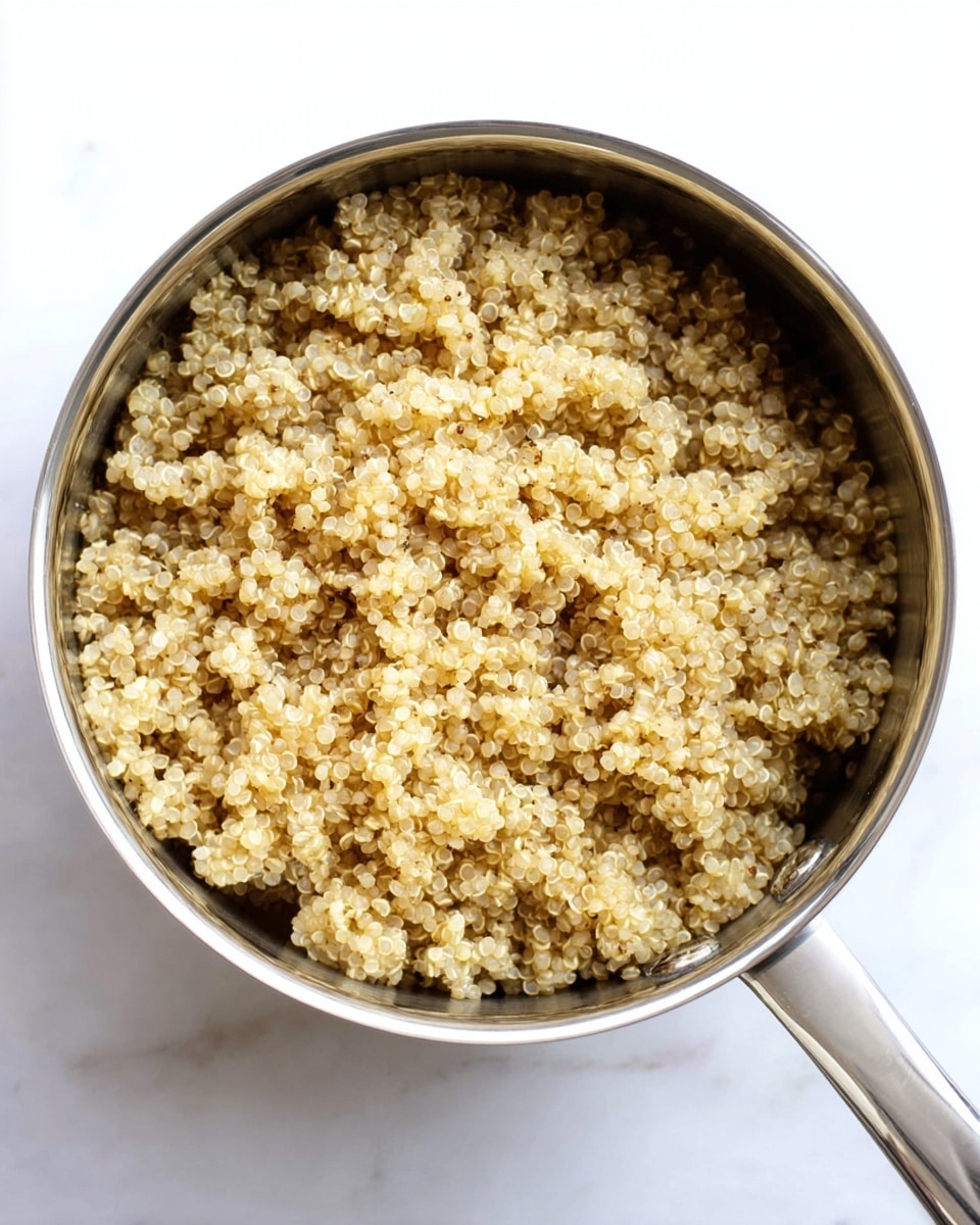 A shiny silver pot is filled with cooked quinoa, showing many small, round grains with a slightly translucent, pale yellow color. The quinoa has a soft, fluffy texture with tiny curly tails visible on each grain. The pot handle extends downward and the whole setup sits on a white marbled surface. photo taken with an iphone --ar 4:5 --v 7