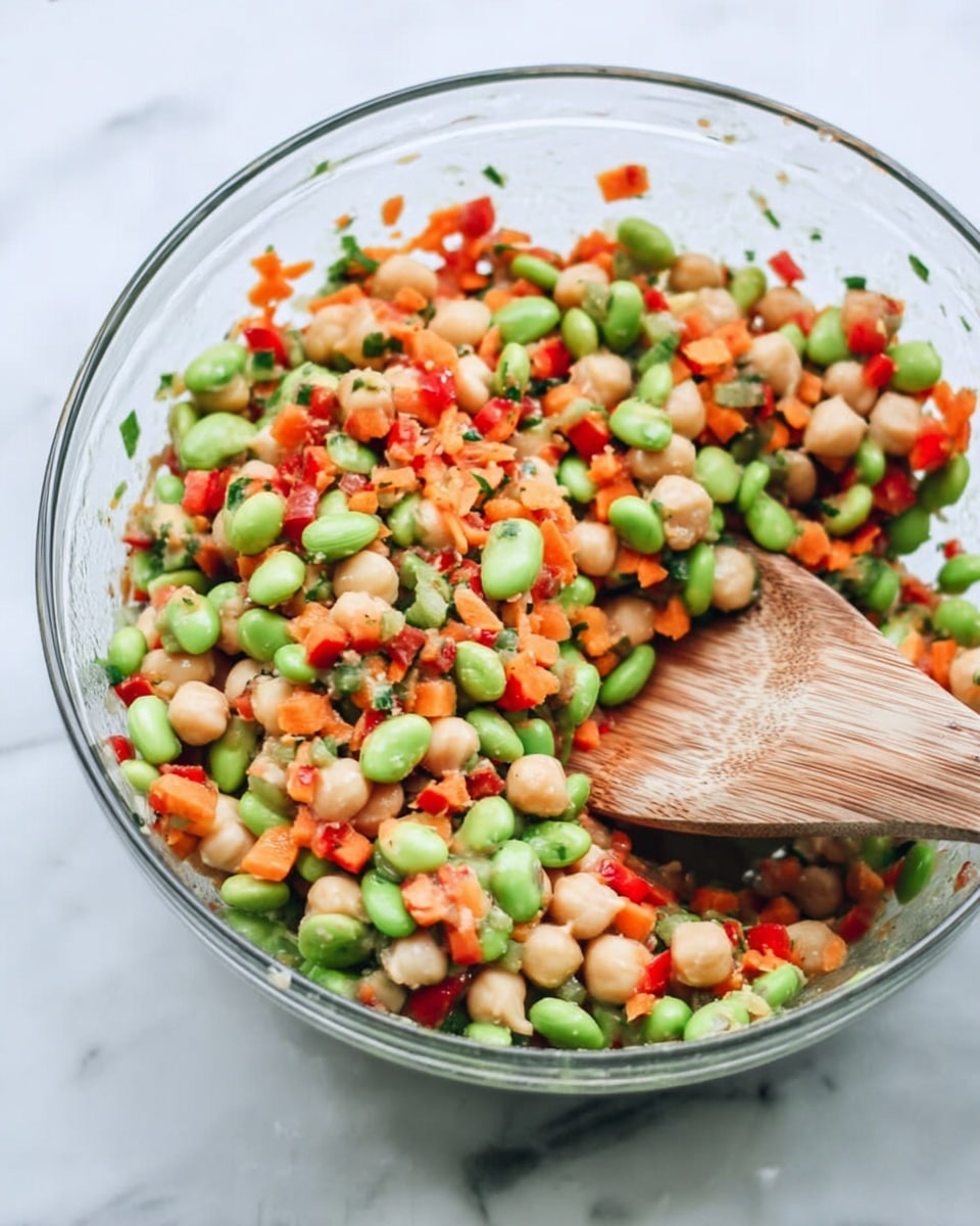The image shows a clear glass bowl filled with a colorful salad made of three main layers: green edamame beans, pale beige chickpeas, and small diced red bell peppers mixed with finely chopped green herbs and orange carrots. The salad has a fresh, mixed texture with all ingredients evenly spread throughout the bowl. A wooden spoon rests inside the bowl, partly covered by the salad, and the background is a white marbled surface. photo taken with an iphone --ar 4:5 --v 7