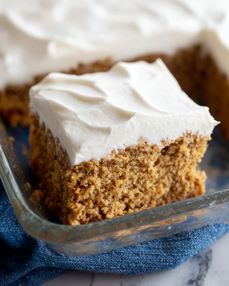 A close-up view of a square piece of cake with two visible layers: the bottom layer is a light brown, moist, and crumbly textured cake, and the top layer is a thick, smooth white frosting spread evenly with soft, wavy ridges. The cake sits in a clear glass baking dish on a white marbled surface with a blue cloth partially visible beneath the dish. The focus is on the front cake piece, showing detailed texture of the crumb and frosting. photo taken with an iphone --ar 4:5 --v 7