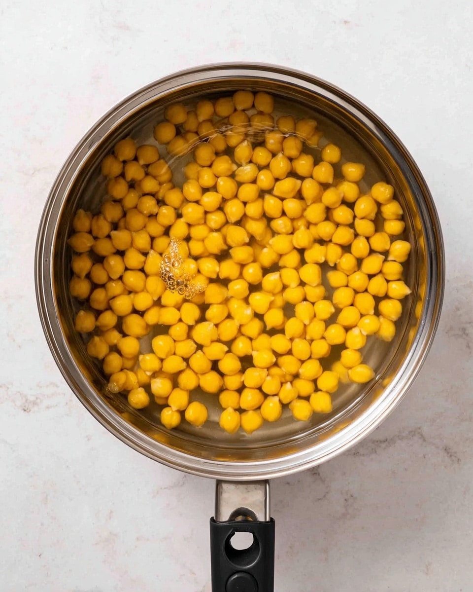 A top-down view of a metal pot filled with water and yellow chickpeas soaking inside. The chickpeas are round and smooth, densely packed but with some space between each, visible under the clear water. The pot has a black handle on one side and a black grip-like handle with a hole on the other side. The pot sits on a white marbled surface that contrasts with the hand. photo taken with an iphone --ar 4:5 --v 7