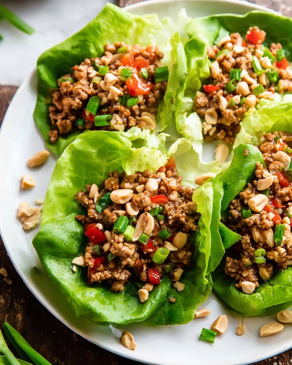 A white plate holds four large bright green lettuce leaves as the first layer, each leaf shaped like a bowl. Inside each leaf is a mix of brown cooked ground meat with small pieces of red bell pepper and onion, forming the second layer. The top is scattered with chopped light beige peanuts and small pieces of green spring onion, adding texture and color contrast. The plate sits on a white marbled surface with some loose chopped spring onion pieces visible around it. Photo taken with an iphone --ar 4:5 --v 7