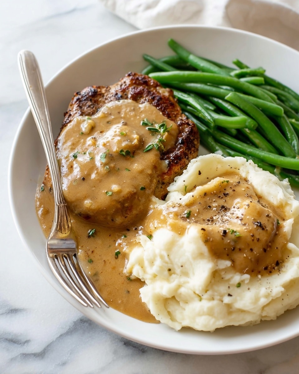 A white plate holds a meal with three parts: on the left, a grilled piece of meat covered by a thick, creamy brown gravy with visible herbs; on the top right, a small pile of bright green steamed green beans arranged neatly; on the bottom right, a large scoop of smooth white mashed potatoes topped with the same brown gravy and sprinkled with black pepper. A silver fork rests on the mashed potatoes, all placed on a white marbled surface. photo taken with an iphone --ar 4:5 --v 7
