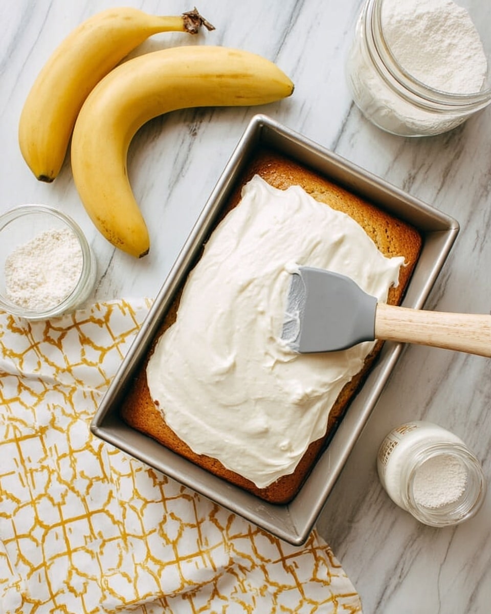 A rectangular golden brown cake sits in a silver baking pan on a white marbled surface. A thick layer of white creamy frosting is being spread on the cake with a gray spatula. To the left of the pan, two yellow bananas rest on a white cloth with a gold geometric pattern. Around the cake pan, there are small kitchen items including a white jar with a lid and a glass container filled with white powder. photo taken with an iphone --ar 4:5 --v 7