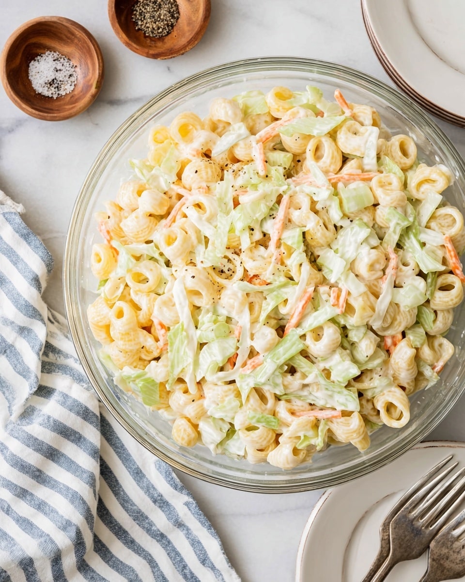 A clear glass bowl filled with a mixed salad that has small pale yellow pasta rings, thin white cabbage strips, light green celery pieces, and thin orange carrot strips, all mixed together with a creamy white dressing. Two wooden small bowls nearby contain black pepper and salt. A neatly folded white cloth with blue stripes is placed at the bottom left of the image, and two white plates stacked with two silver forks rest at the bottom right, all set on a white marbled surface. Photo taken with an iphone --ar 4:5 --v 7