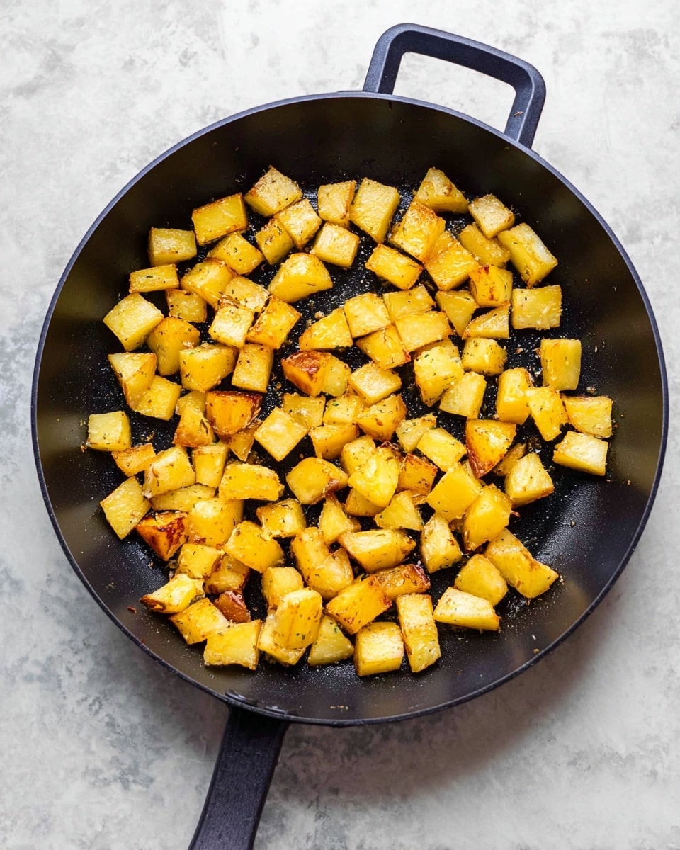 A black pan with a handle filled with small, evenly sized cubes of golden brown cooked potatoes spread out inside. The potato cubes have a slight shine and some light seasoning visible on their surface, with a mix of soft yellow and light brown colors. The pan is placed on a white marbled surface. photo taken with an iphone --ar 4:5 --v 7