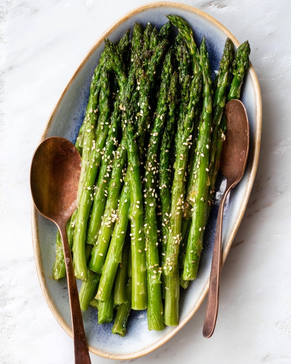 A white oval plate holds a neat pile of bright green roasted asparagus spears, arranged in one layer with their tips all pointing in the same direction at the top. The asparagus shows slight charring and shine from oil, sprinkled with small white sesame seeds scattered evenly on top. Two vintage copper salad spoons rest under the asparagus on either side, their round, worn surfaces visible. The plate is placed on a white marbled surface that adds a clean, fresh feel to the image. photo taken with an iphone --ar 4:5 --v 7