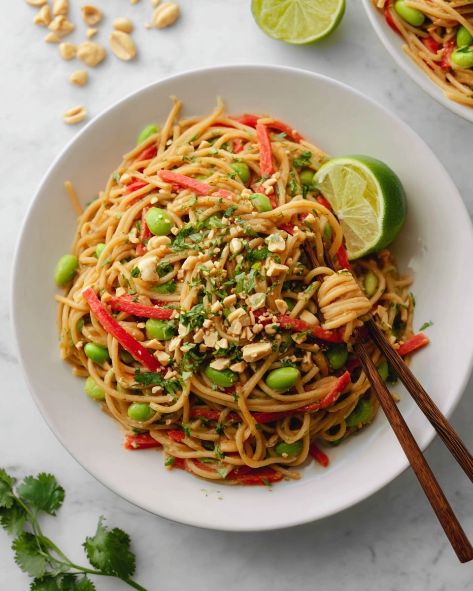The dish shows a white round plate filled with light brown noodles mixed with bright green edamame beans, thin red bell pepper strips, and small pieces of chopped peanuts scattered on top. Fresh green herbs are sprinkled over the noodles, adding a fresh touch. On the side of the noodles in the plate, a green lime wedge rests against the noodles. Wooden chopsticks are placed on the right side, partially holding the noodles. In the background, there is a white marbled surface with half a lime visible above the plate and some peanuts and green herbs arranged around the plate. photo taken with an iphone --ar 4:5 --v 7