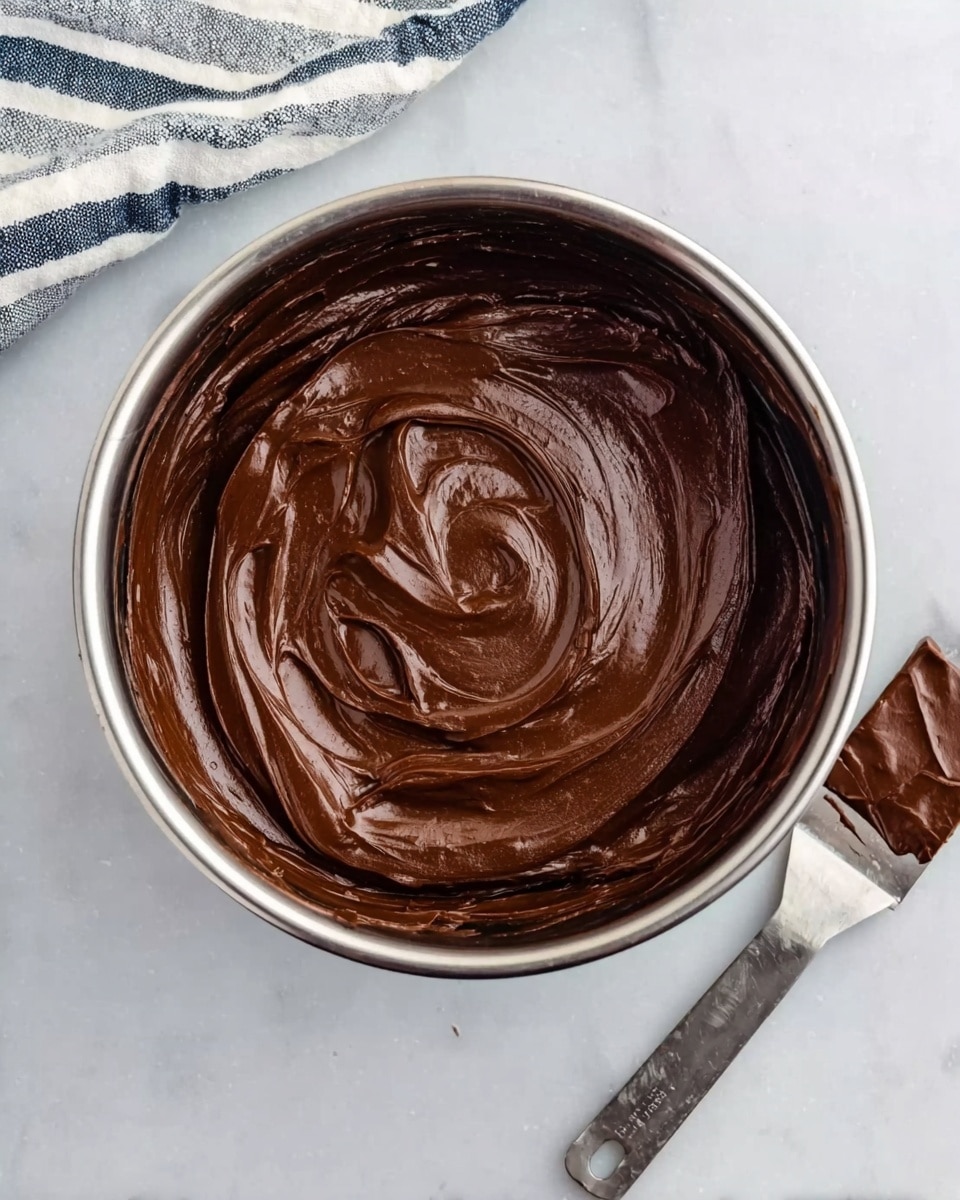 The image shows a shiny metal pot filled with smooth dark brown chocolate spread with thick, creamy texture. The chocolate is swirled in the pot with visible curves and folds on the surface, making it look soft and rich. The pot is placed on a white marbled surface, and near the pot, on the right side, there is a small metal spatula covered in the same dark chocolate. A striped cloth is partially visible in the top left corner, adding a bit of color contrast. photo taken with an iphone --ar 4:5 --v 7