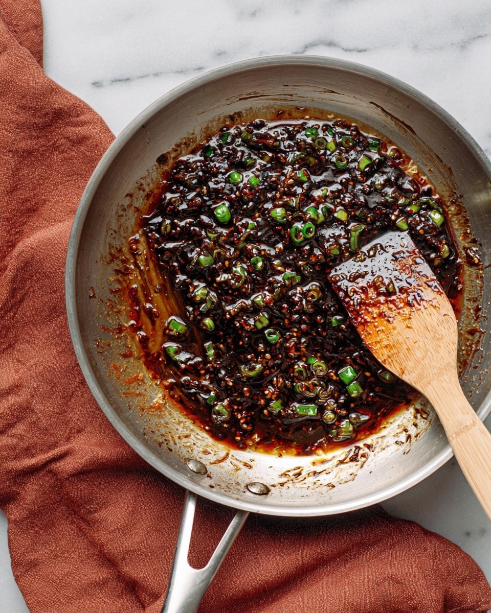 In a shiny silver pan, there is a thick, dark brown sauce mixed with small green pieces of chopped spring onions spread mainly in the center and some smudges on the sides. The sauce has a glossy texture with some oily red highlights. On the right side inside the pan, a wooden spatula is partially covered with the sauce. The pan rests on a soft, wrinkled rusty red cloth, and the background shows a white marbled surface. photo taken with an iphone --ar 4:5 --v 7