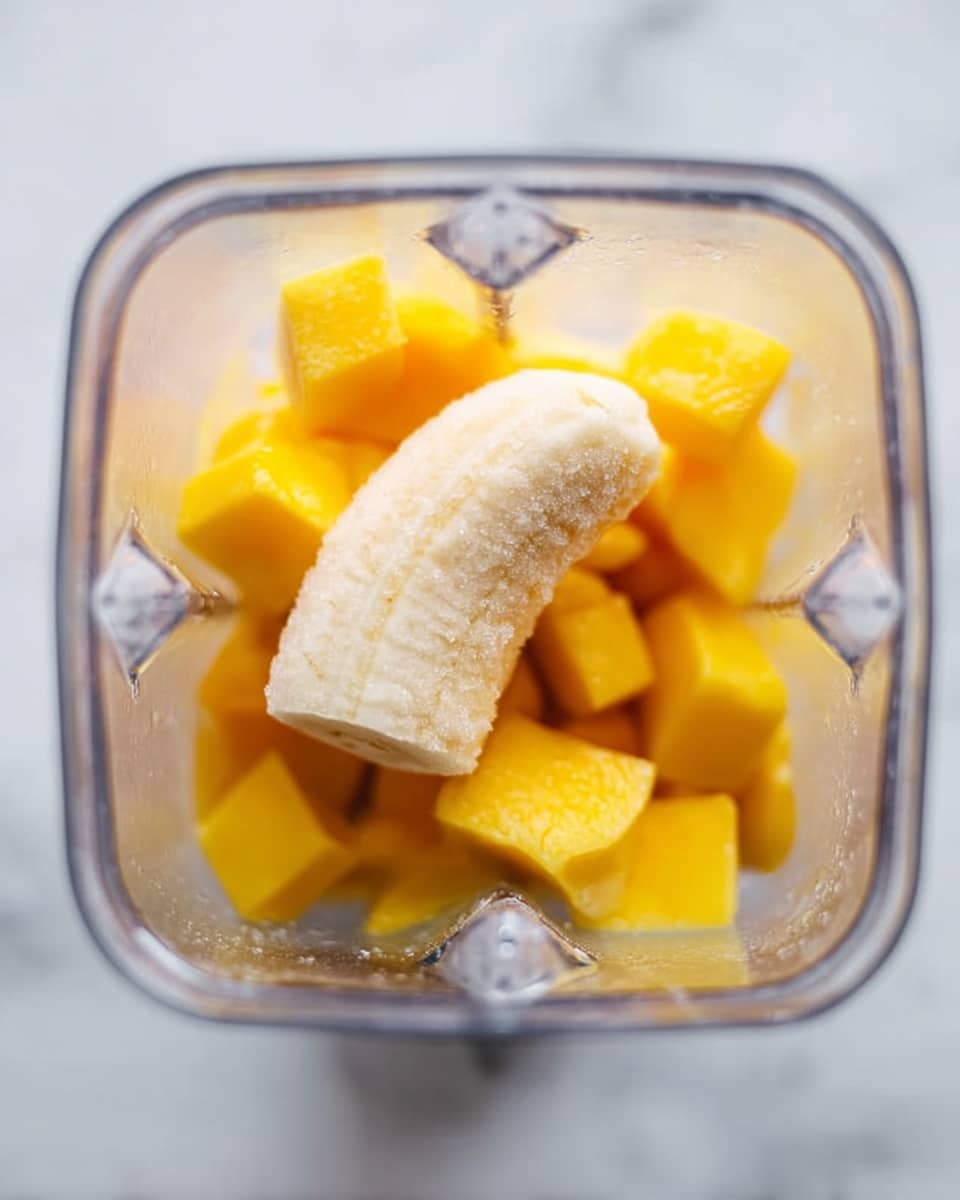 The image shows a close-up top view inside a transparent blender container. The blender holds one peeled frozen banana piece that is light cream in color with a slightly frosty texture, placed in the center. Around the banana, there are bright yellow frozen mango cubes scattered evenly, with smooth icy surfaces. The background outside the blender has a white marbled texture. photo taken with an iphone --ar 4:5 --v 7