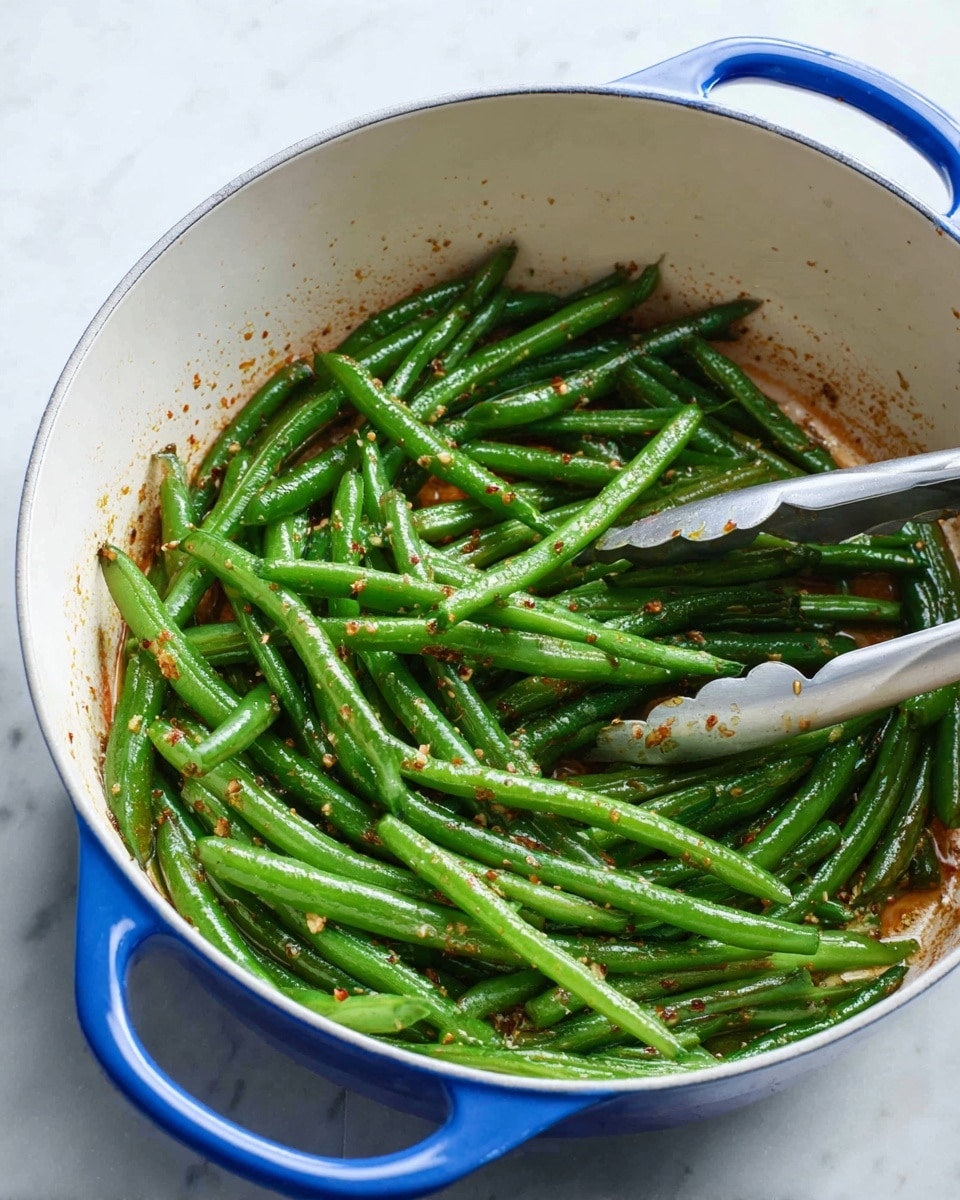 Inside a white cooking pot with blue handles, there are cooked green beans spread on one layer. The green beans are shiny and bright green with some light brown seasoning bits on top. A pair of silver tongs grasps some green beans on the right side. The pot sits on a white marbled surface. photo taken with an iphone --ar 4:5 --v 7