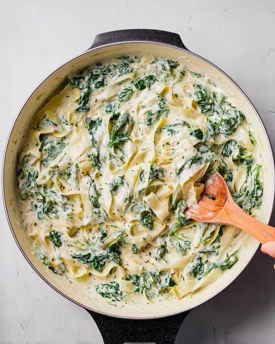 A large white pan filled with a creamy sauce mixed with bright green spinach leaves and wide flat pasta strips. The sauce is thick and white, covering the pasta and spinach evenly. A woman's hand holds a wooden spoon stirring the mixture gently from the right side. The background is a white marbled surface, adding brightness to the scene. photo taken with an iphone --ar 4:5 --v 7