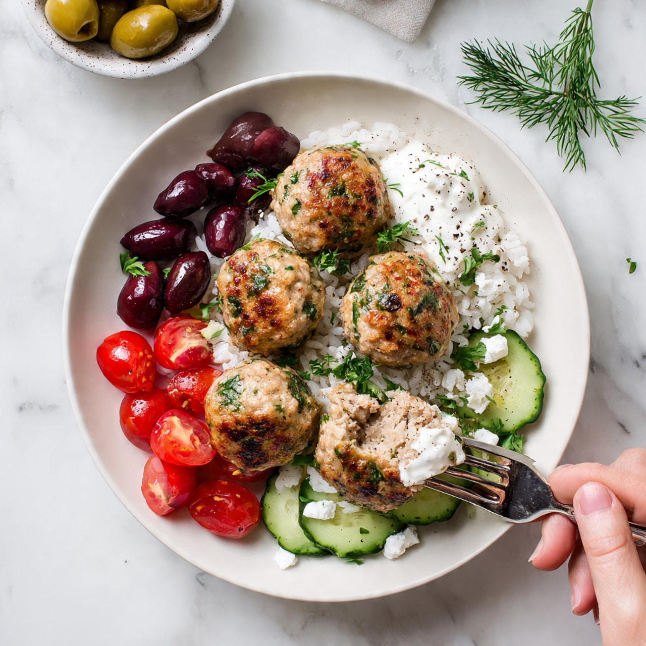 A white plate holds a colorful dish with five light brown meatballs with green herbs mixed in, placed on a bed of white rice. The meatballs are arranged near the center with a woman's hand holding a fork that has poked one meatball, showing its inside texture. Around the meatballs, there are layers of sliced dark purple olives on the left, bright red cherry tomatoes in the front with small white cheese crumbles, and light green cucumber slices on the right next to a dollop of white sauce sprinkled with green herbs and black pepper. Fresh dill and parsley leaves are scattered over the dish. The plate is set on a white marbled surface with two small bowls partially visible at the top edge, one filled with olives. Photo taken with an iphone --ar 4:5 --v 7