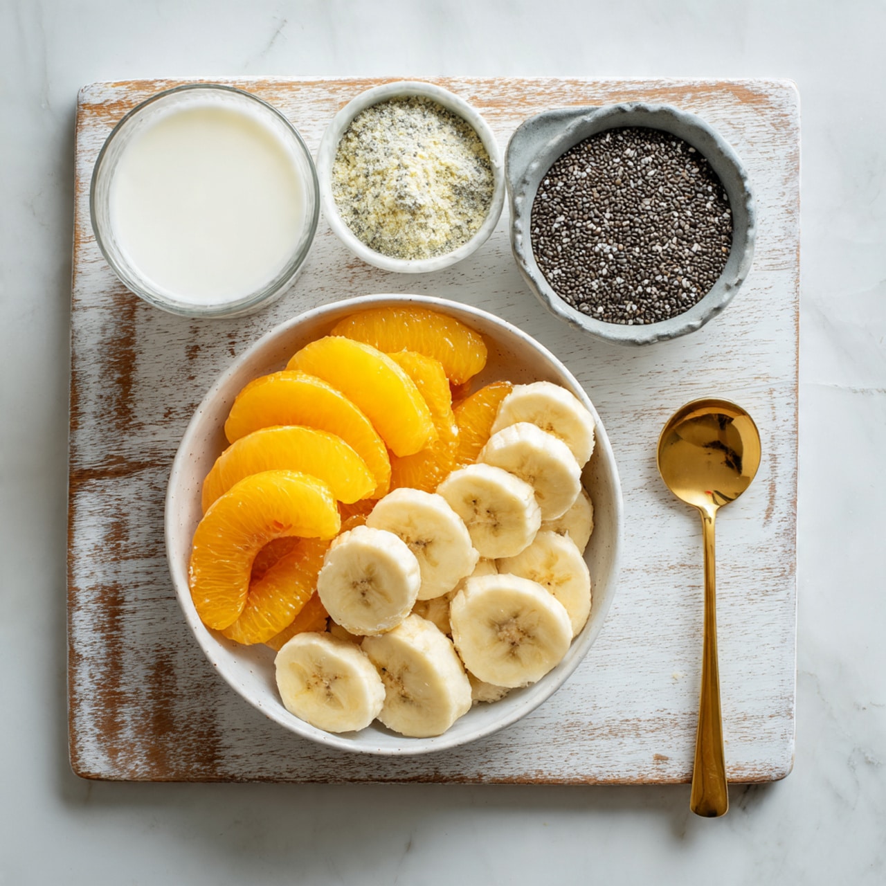 A white bowl filled with two layers of sliced fruits, the bottom layer is orange peach slices with smooth edges, and the top layer is pale yellow banana slices stacked neatly on one side. To the right of the bowl, there is a gray measuring cup filled with small black chia seeds that have a textured surface. Below the cup, a smaller white bowl contains off-white powder with some clumps and a hint of green on the side. To the left, a clear glass filled with white liquid sits on a white marbled surface, and next to it lies a shiny golden spoon with a round head. The entire setup is placed on a well-used white wooden board with visible scratches and some light brown marks. Photo taken with an iphone --ar 4:5 --v 7