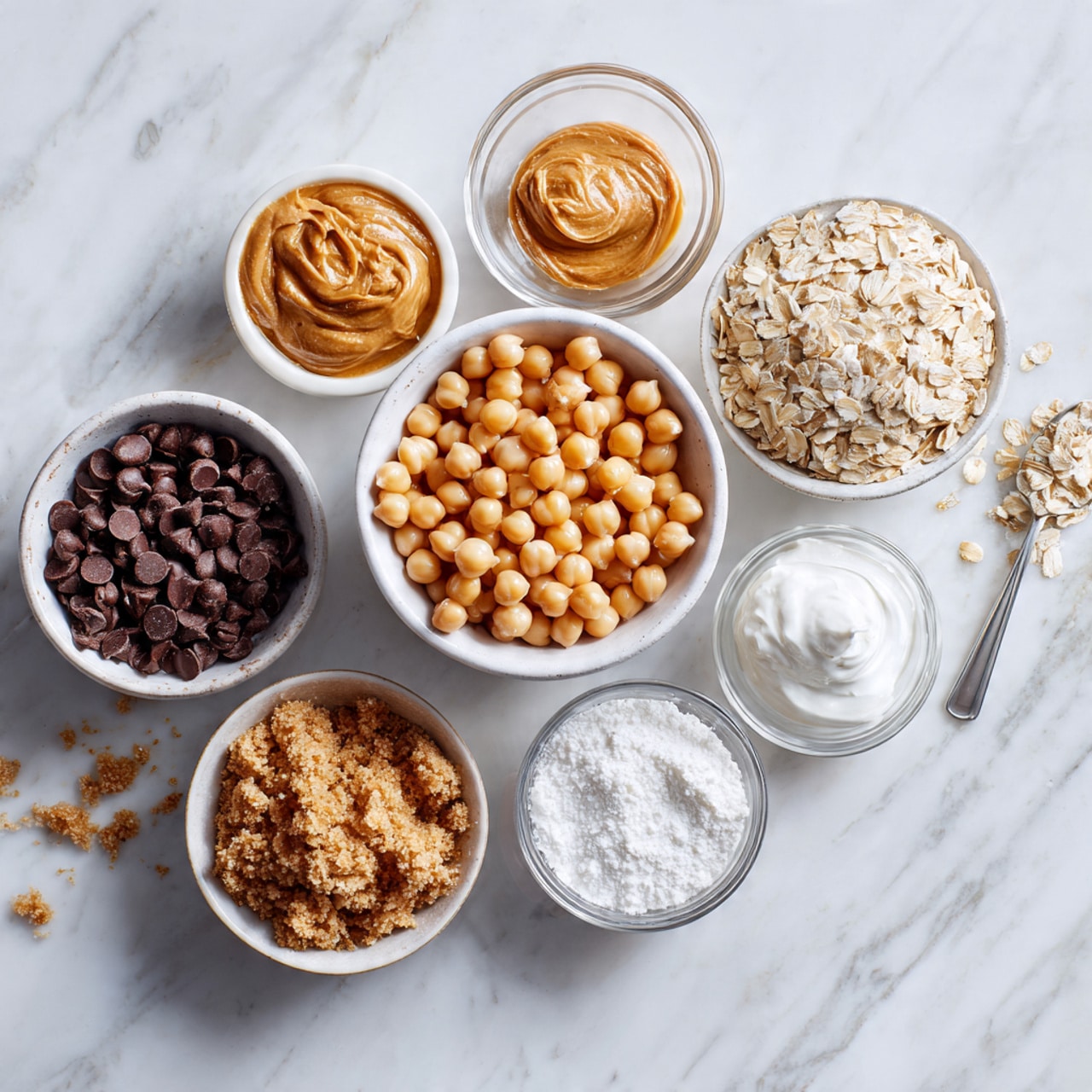 The image shows seven bowls arranged on a white marbled surface. In the center is a white bowl full of light beige chickpeas, round and smooth, filling the bowl nearly to the top. Surrounding it are six smaller white bowls: one has dark brown chocolate chips, shiny and rounded; another holds tan peanut butter with a creamy texture; a third has light beige rolled oats with a dry, flaky look; one is filled with light brown crumbly brown sugar; another contains shiny white yogurt with creamy peaks; and the last has white powdery baking soda. Behind the bowls is a small empty clear glass bowl. A woman's hand holding a small spoon is reaching from the right side near the chocolate chips. photo taken with an iphone --ar 4:5 --v 7