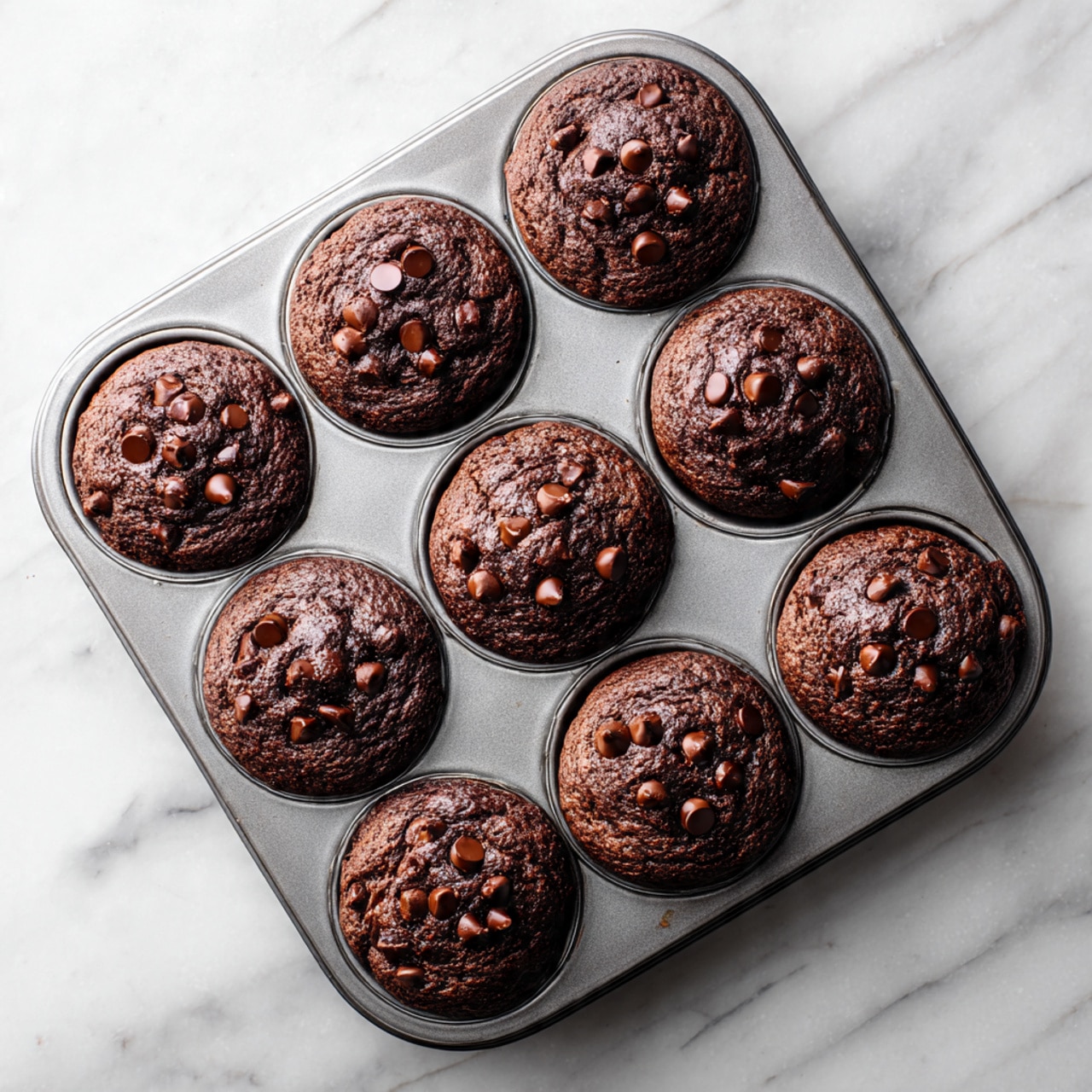 The image shows five chocolate muffins placed closely together on a white plate. Each muffin has a dark brown color with a slightly rough, cracked top, sprinkled with small shiny chocolate chips. The muffins are wrapped in dark purple paper liners that contrast with the chocolate tops. The plate sits on a white marbled textured surface, creating a clean and bright background. The focus is sharp on the muffins in front, with a soft blur on the ones behind them, showing depth. photo taken with an iphone --ar 4:5 --v 7