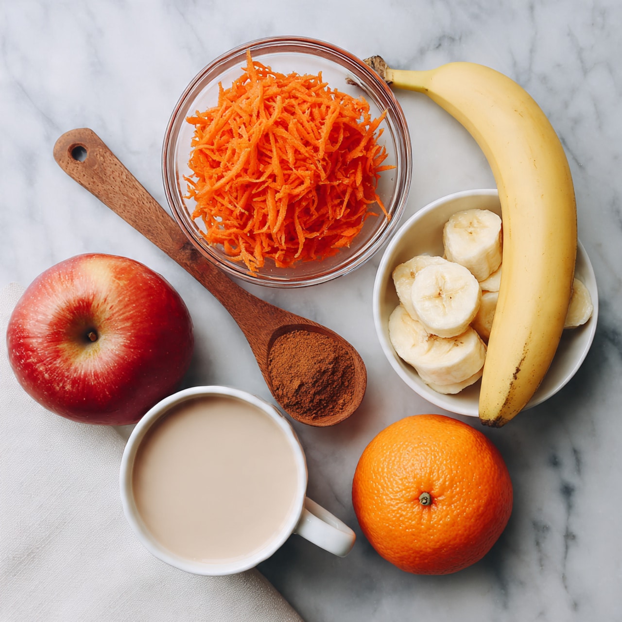 A white marbled surface holds several food items: a glass bowl with bright orange shredded carrots at the top center, a wooden spoon below it filled with ground cinnamon, a white bowl on the right filled with sliced yellow bananas, a whole bright orange fruit on the left, a red apple with a small stem beside the orange, and a glass cup with a creamy light beige liquid at the bottom center. A white cloth is slightly visible on the lower left corner. The scene looks fresh and simple, with natural colors and soft light. photo taken with an iphone --ar 4:5 --v 7