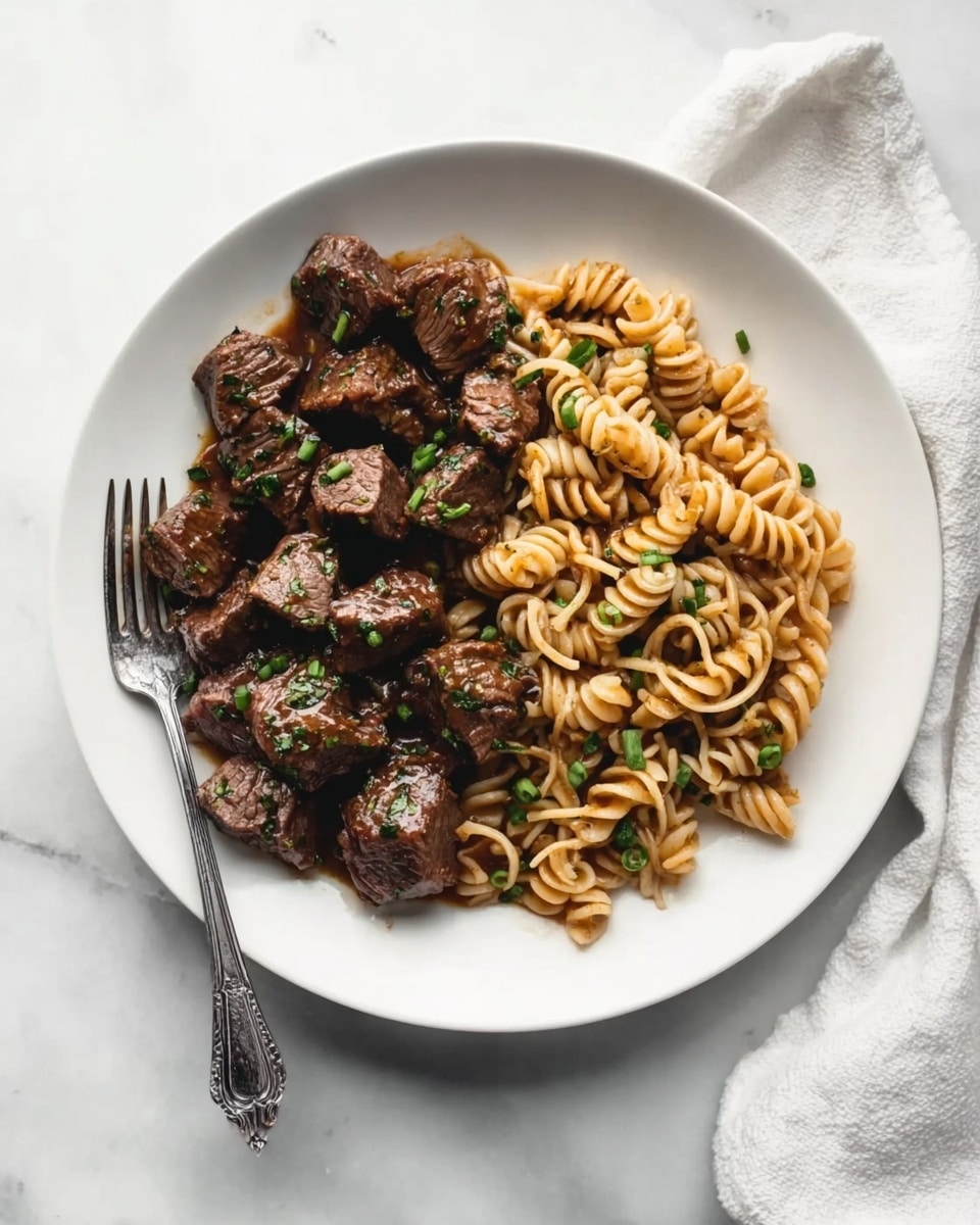 The dish shows a white round plate on a white marbled surface with cooked beef cubes arranged on the left side of the plate. The beef is dark brown with a slight shine, and has some green herbs sprinkled on top. On the right side, there is a pile of spiral-shaped noodles mixed with small green vegetable pieces, all lightly browned and glossy. A silver fork rests on the left edge of the plate, with its tines pointing towards the beef. A white cloth napkin is partly visible in the top right corner of the image. Photo taken with an iphone --ar 4:5 --v 7