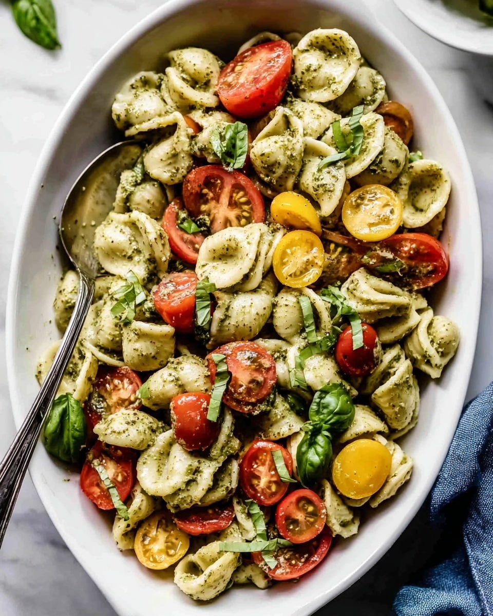 A white oval bowl sits on a white marbled surface filled with a colorful pasta salad. The salad has three layers: the base is made of round, shell-shaped pasta coated in a green pesto sauce, showing a slightly rough texture. Mixed throughout are sliced red and yellow cherry tomatoes, adding bright pops of color with their smooth, shiny skins. Scattered on top are fresh green basil leaves that add a fresh touch. A silver spoon rests inside the bowl on the left side, slightly submerged in the pasta, and a corner of a blue cloth is visible on the bottom right. Photo taken with an iphone --ar 4:5 --v 7