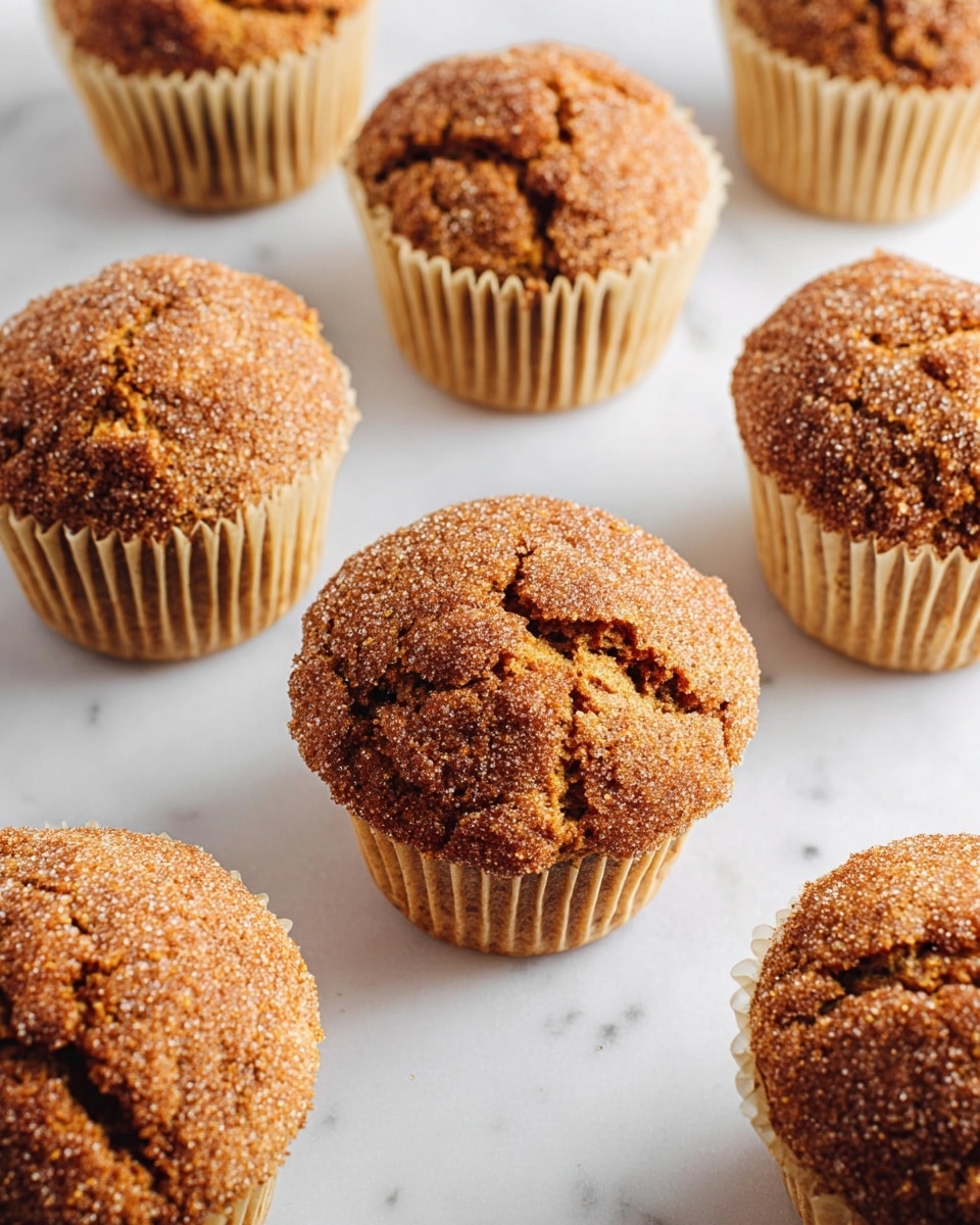 The image shows eight round muffins with a rough, crumbly top layer in a golden brown color, sprinkled with small granules that add texture. Each muffin is held in a pale beige paper liner, and the muffins have cracks and uneven surfaces which give a rustic look. The muffins are arranged evenly on a surface with a white marbled texture, with soft natural light highlighting the warm tones of the muffin tops. The focus is sharp on the muffin closest to the center while others gently blur towards the edges. photo taken with an iphone --ar 4:5 --v 7