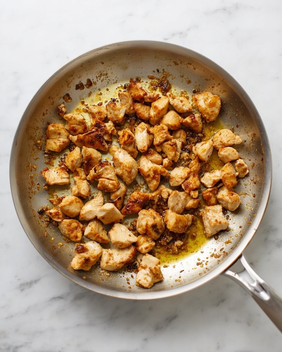 A silver metal frying pan filled with small pieces of cooked chicken that are golden brown and slightly crispy. The chicken pieces are spread unevenly across the pan's inside, with some bits showing darker, almost caramelized edges. The pan rests on a white marbled surface, and the cooked chicken sits in a thin layer of light oil and browned bits, giving a textured look to the whole scene. Photo taken with an iphone --ar 4:5 --v 7