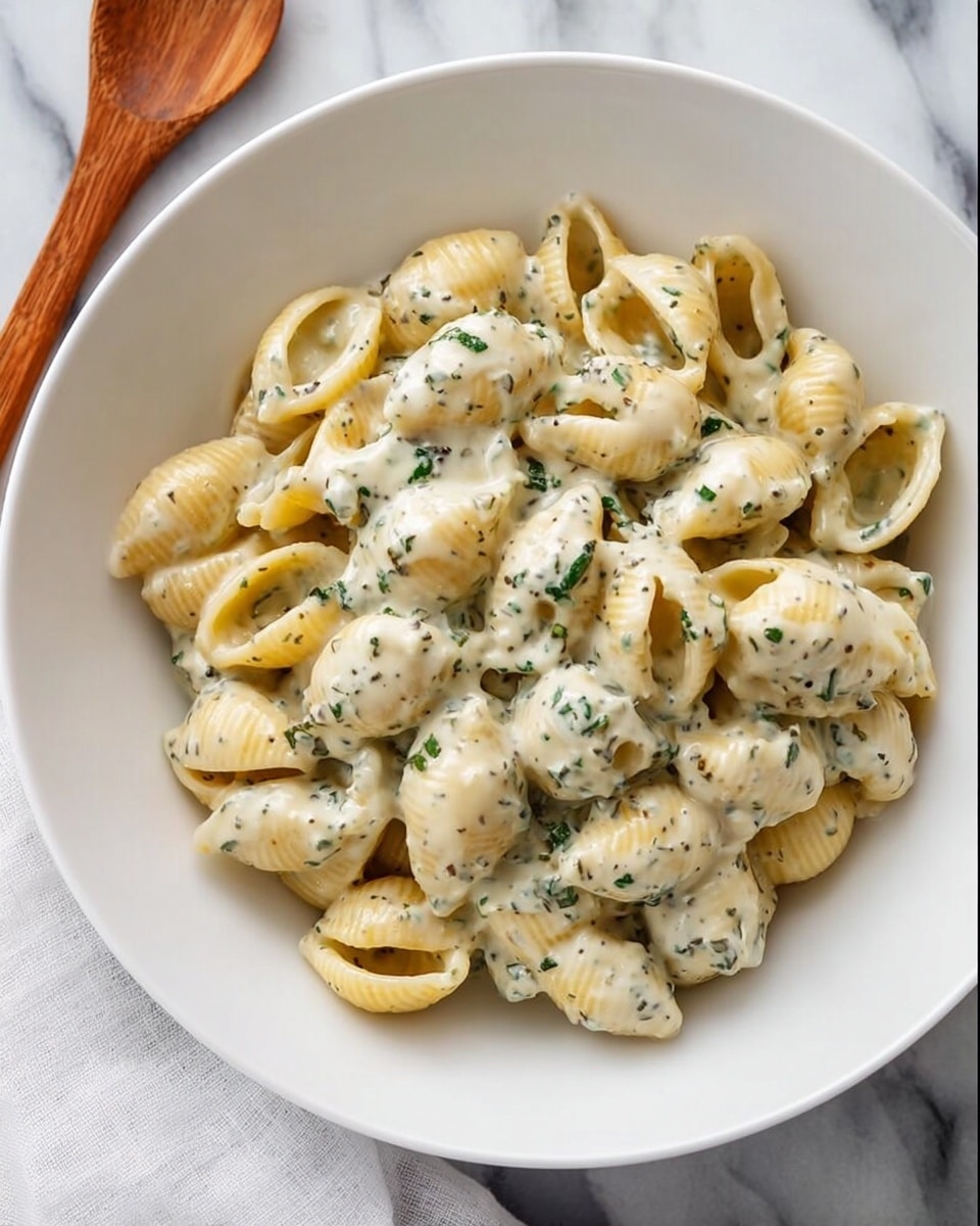 A white bowl filled with a single layer of shell pasta covered evenly with creamy white sauce speckled with dark green herbs, the sauce giving a smooth and rich texture that clings to each pasta piece, all placed on a white marbled surface with a folded white cloth napkin nearby, photo taken with an iphone --ar 4:5 --v 7