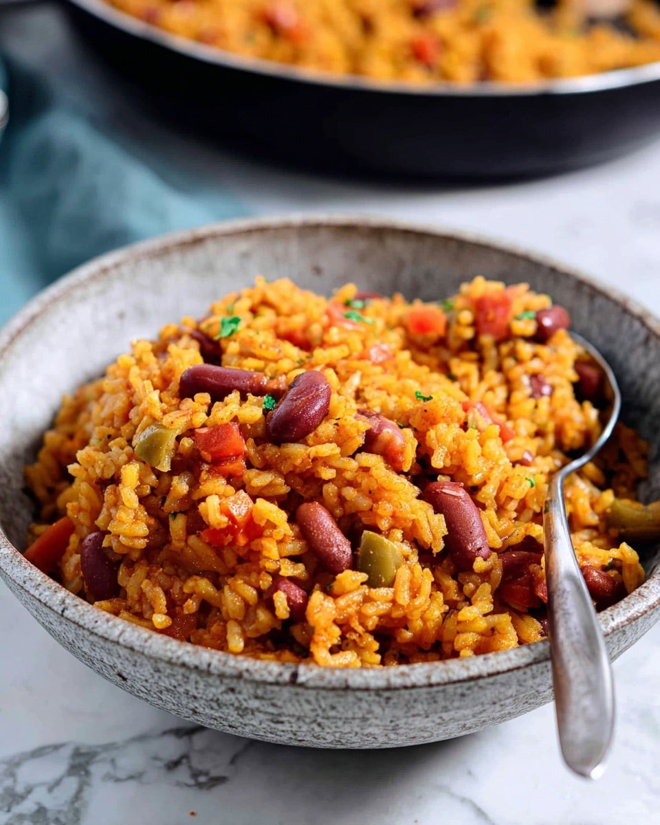 A close-up of a gray speckled bowl filled with a mix of cooked yellow-orange rice, red kidney beans, and small chunks of red tomatoes and green olives, showing a soft and slightly moist texture. The bowl has a rustic look and contains a silver spoon resting inside the rice mix on the right side. In the background, there is a blurred dark pan with more rice and beans. The whole scene is set on a white marbled surface. photo taken with an iphone --ar 4:5 --v 7