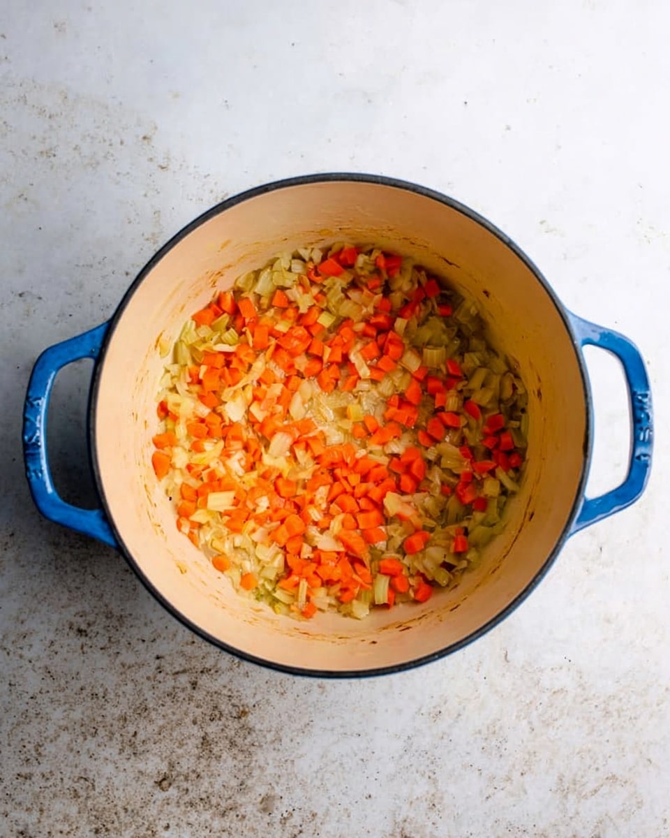A top-down view of a blue enamel pot with two handles on each side, sitting on a white marbled surface. Inside the pot, there is a single layer of diced vegetables consisting mostly of small orange carrot pieces mixed with some beige or light yellow diced onions and celery, all lightly sautéed and spread evenly across the bottom. The inside of the pot has a cream-colored interior with slight browning along the edges where the vegetables meet the pot. photo taken with an iphone --ar 4:5 --v 7