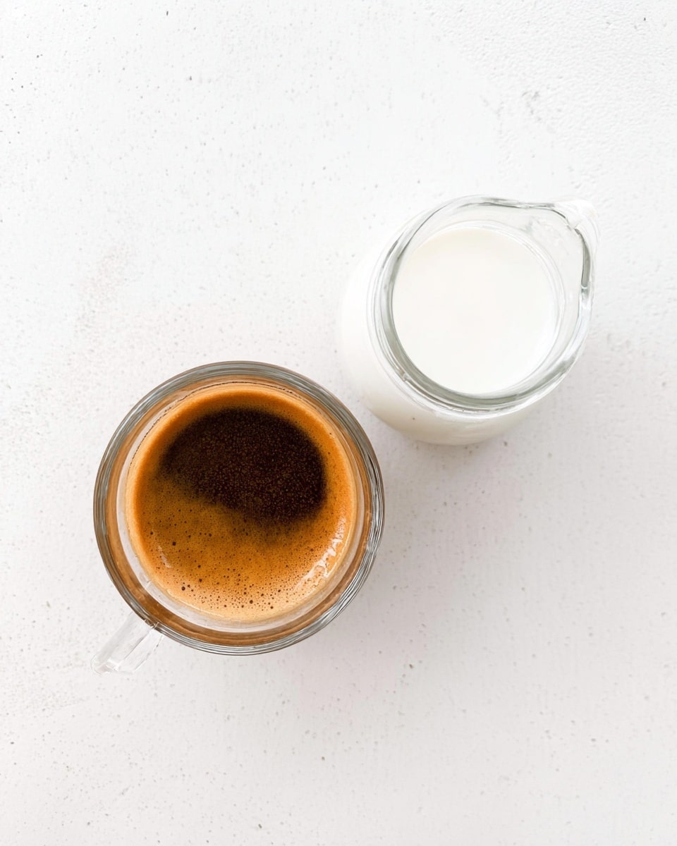 The image shows a small clear glass cup filled with dark brown espresso that has a layer of light brown foam on top. Next to it, there is a small clear glass jug filled with white milk. Both cups are placed on a white marbled surface, creating a clean and simple look. The photo is taken from above, showing the smooth texture of the espresso foam and the shiny surface of the milk jug. photo taken with an iphone --ar 4:5 --v 7
