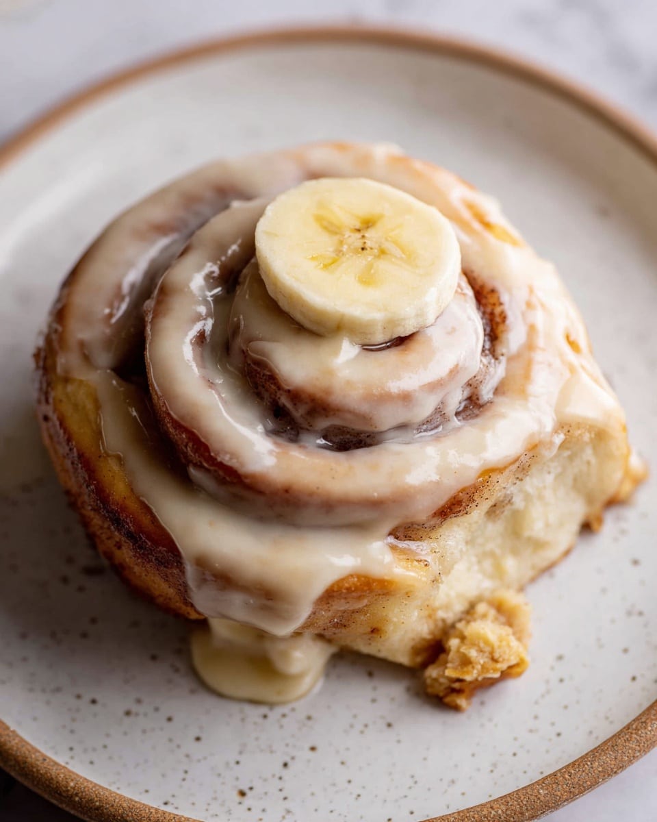 A close-up view of a single cinnamon roll sits centered on a white plate with small gray speckles, placed on a white marbled surface. The cinnamon roll has three visible spiral layers, each showing a golden-brown baked dough with a slightly glossy texture from the baked cinnamon sugar filling. A smooth, creamy beige icing is generously spread over the top and sides of the cinnamon roll, pooling slightly in some areas. On the very top center, there is a single round slice of banana with a light yellow color and faint brown spots. A small piece of crumb sits near the bottom edge of the plate. Photo taken with an iphone --ar 4:5 --v 7